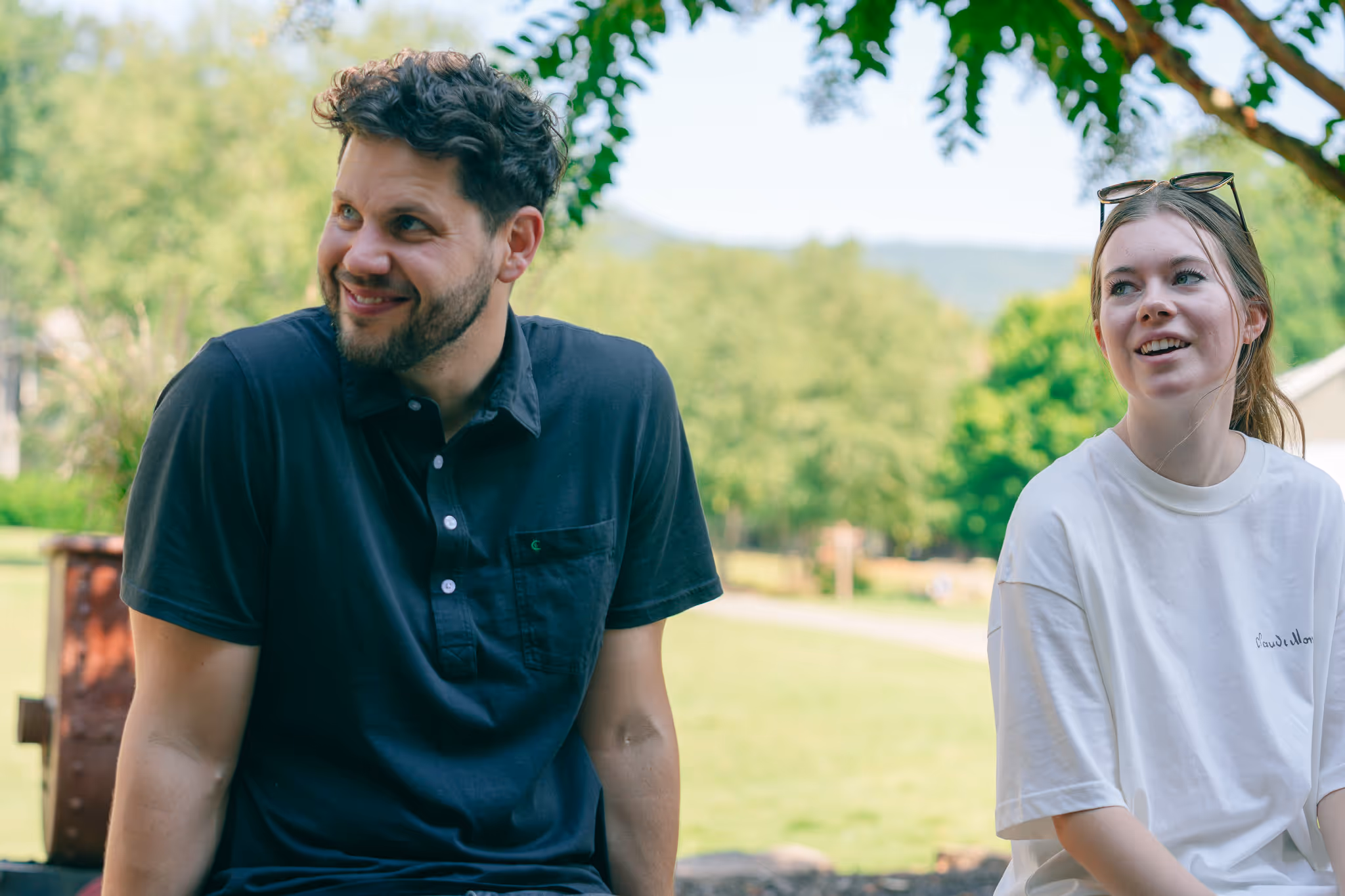 Smiling man in a black shirt and woman in a white shirt with sunglasses on her head sitting outdoors with green trees in the background.