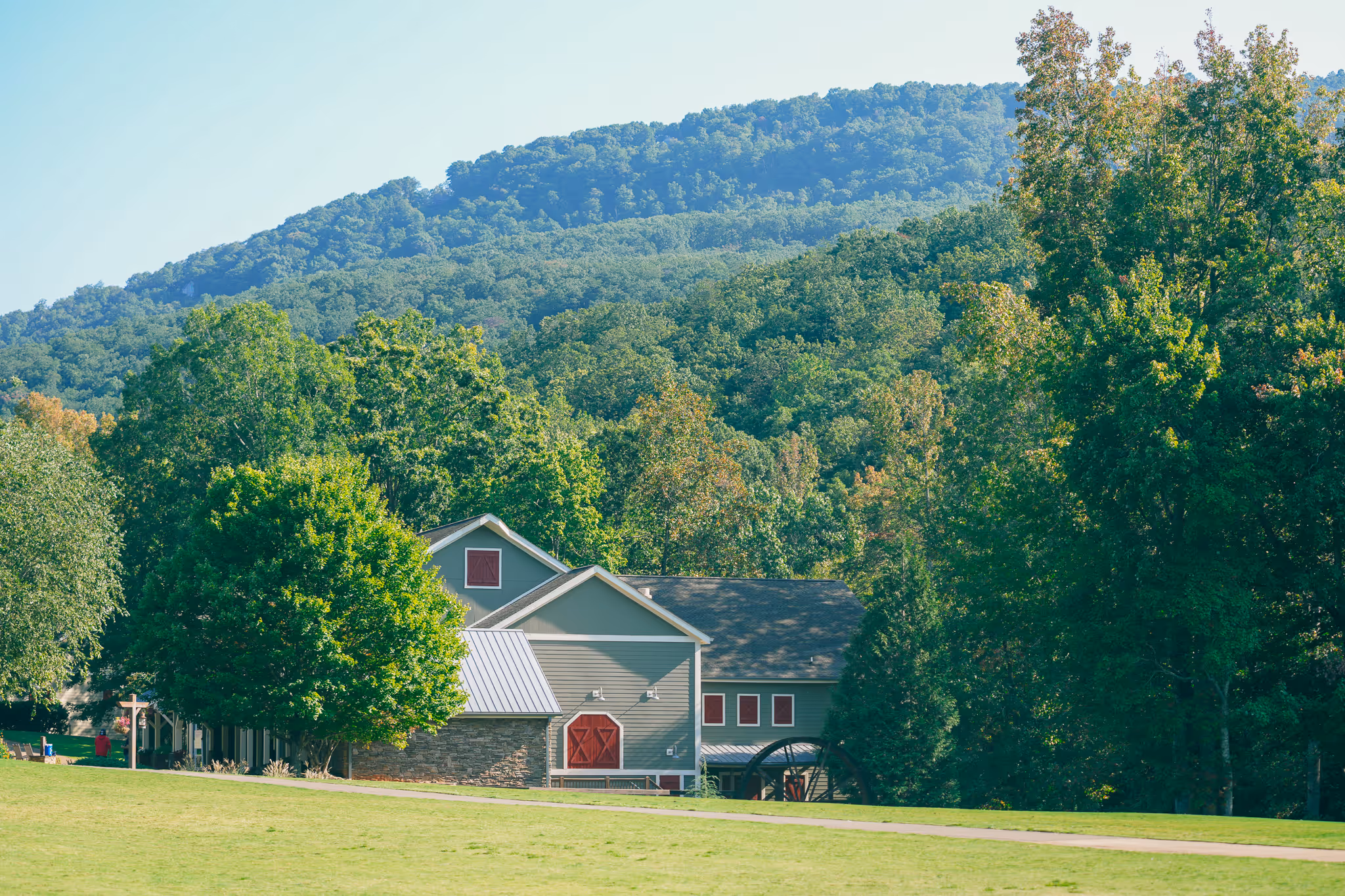 Large green barn with red doors surrounded by trees with forested hills in the background under clear sky.