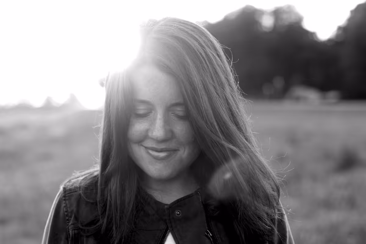 Smiling young woman with long hair and closed eyes in an outdoor field with sunlight behind her.