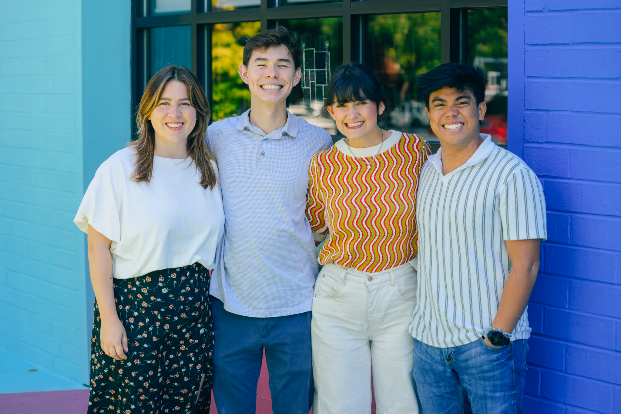 Four young adults smiling and standing arm in arm against a backdrop of turquoise and purple painted brick walls.