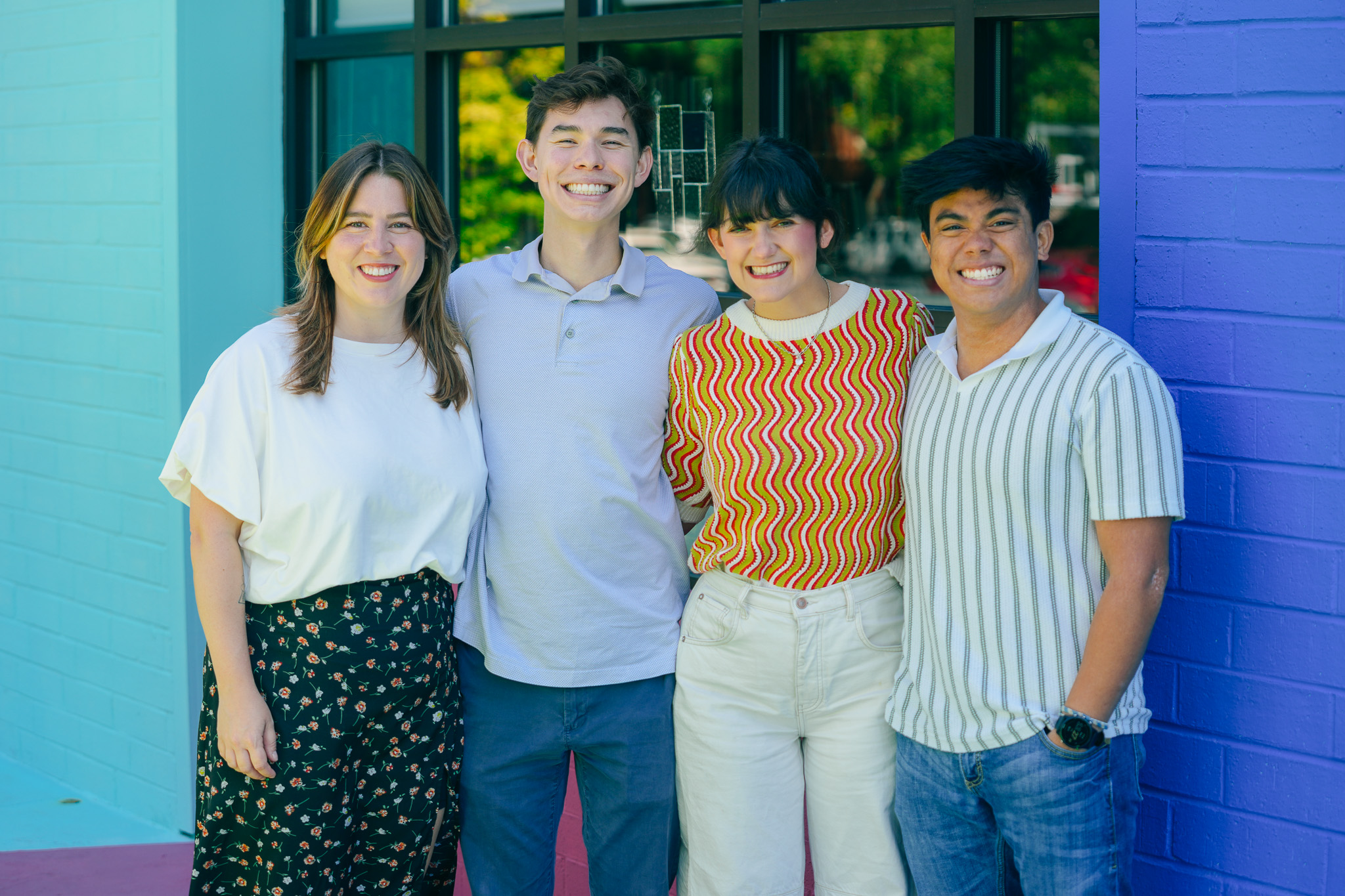 Four young adults smiling and standing arm in arm against a backdrop of turquoise and purple painted brick walls.