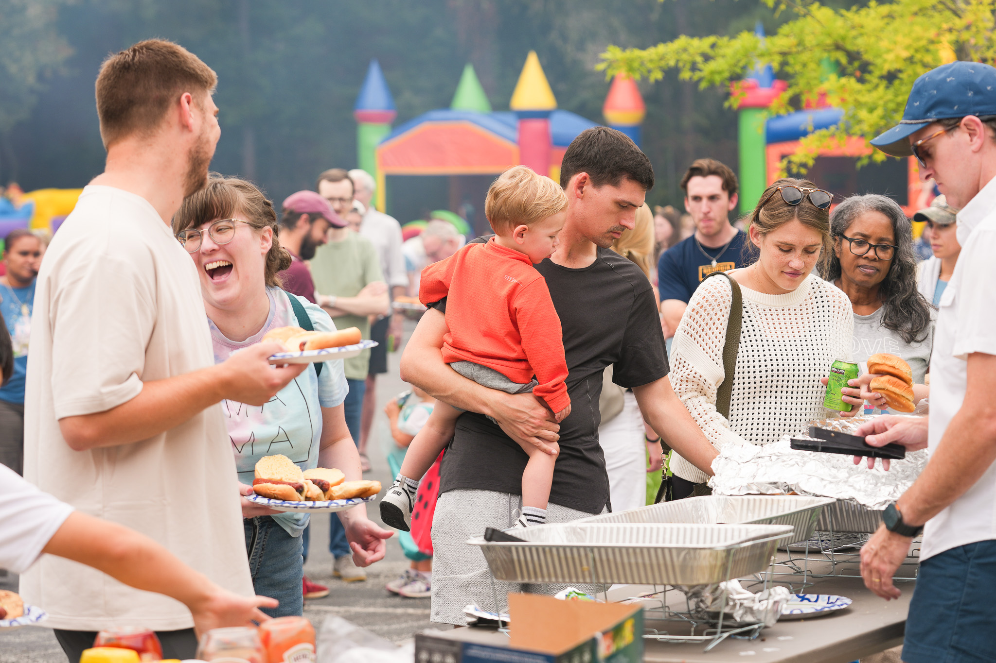 People gathered outdoors around a food table, a man holding a child, and others holding plates with hot dogs at a community event.