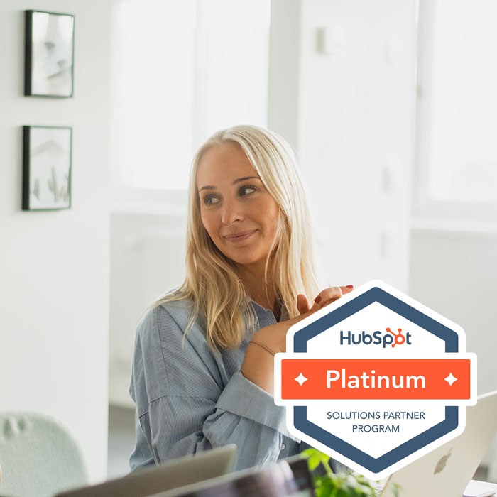 Kvinna med långt blont hår och blå skjorta sitter vid ett bord i ett ljust rum med två inramade tavlor på väggen, med HubSpot Platinum Solutions Partner-programs märke överlagt.