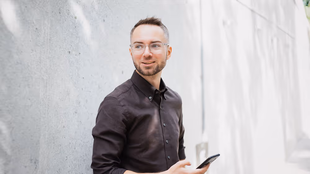 Man med glasögon och skägg håller en smartphone medan han står vid en ljusgrå betongvägg och ser åt sidan.
