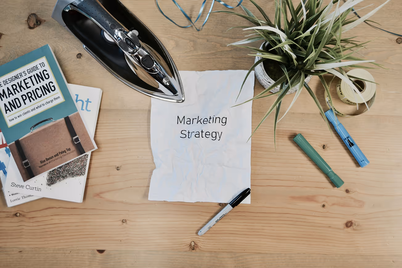 Wooden desk with a crumpled paper reading 'Marketing Strategy,' a hardcover book about marketing and pricing, a black iron, a potted plant, markers, and a roll of tape.