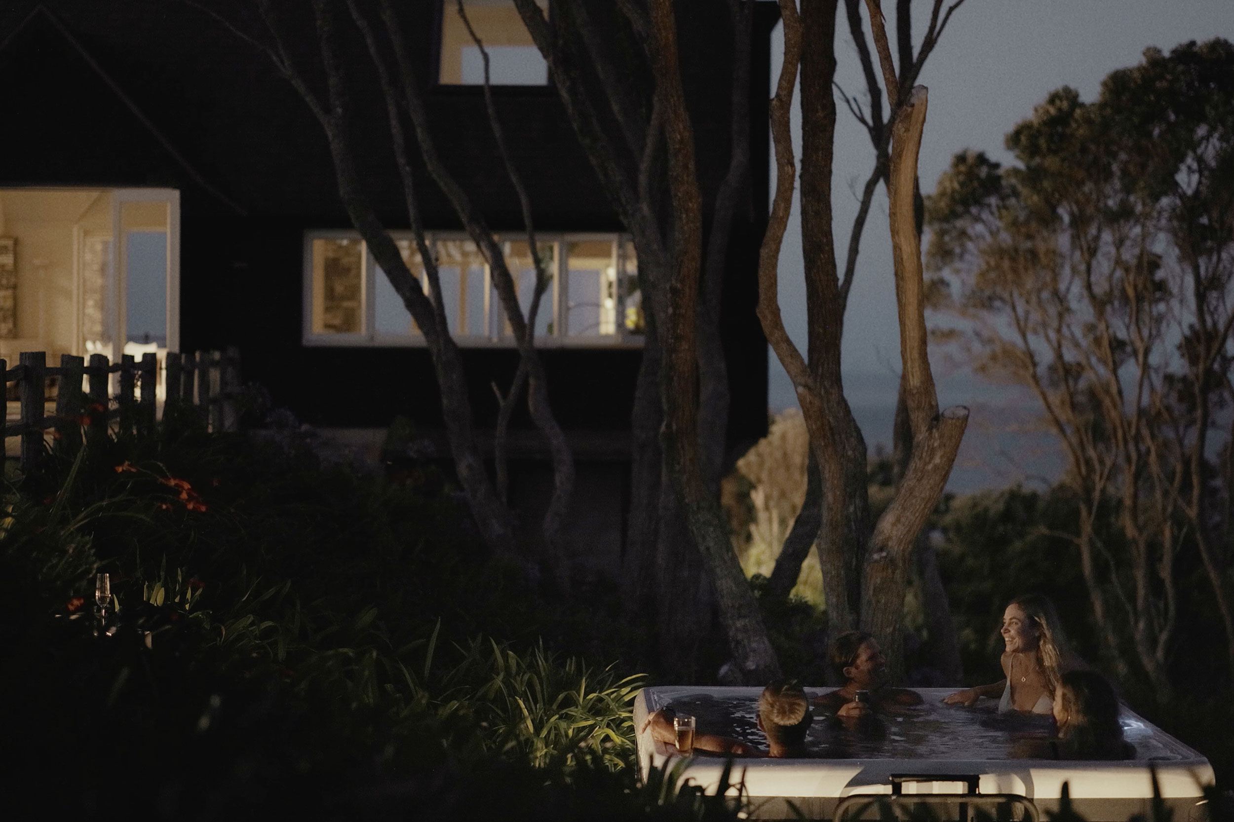 Four people relaxing in an outdoor hot tub at night surrounded by trees and a lit house.