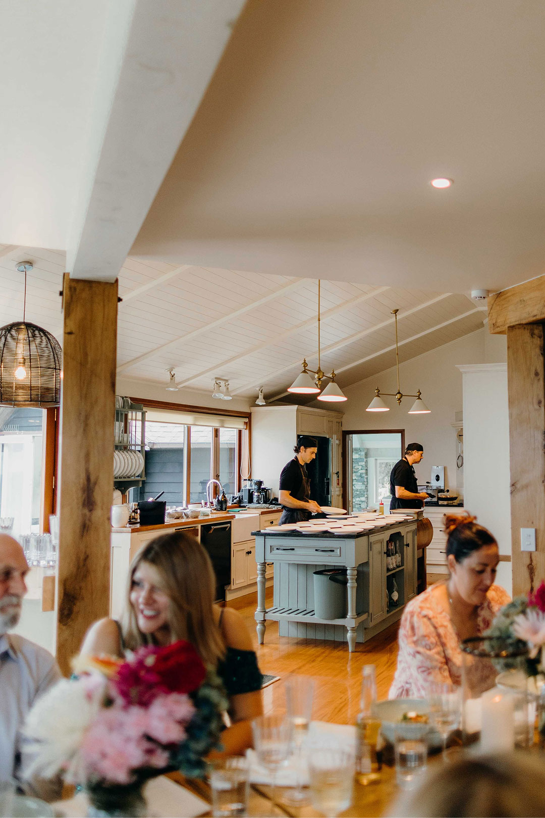 Two chefs preparing plates in a bright kitchen with diners in foreground at a wooden table.