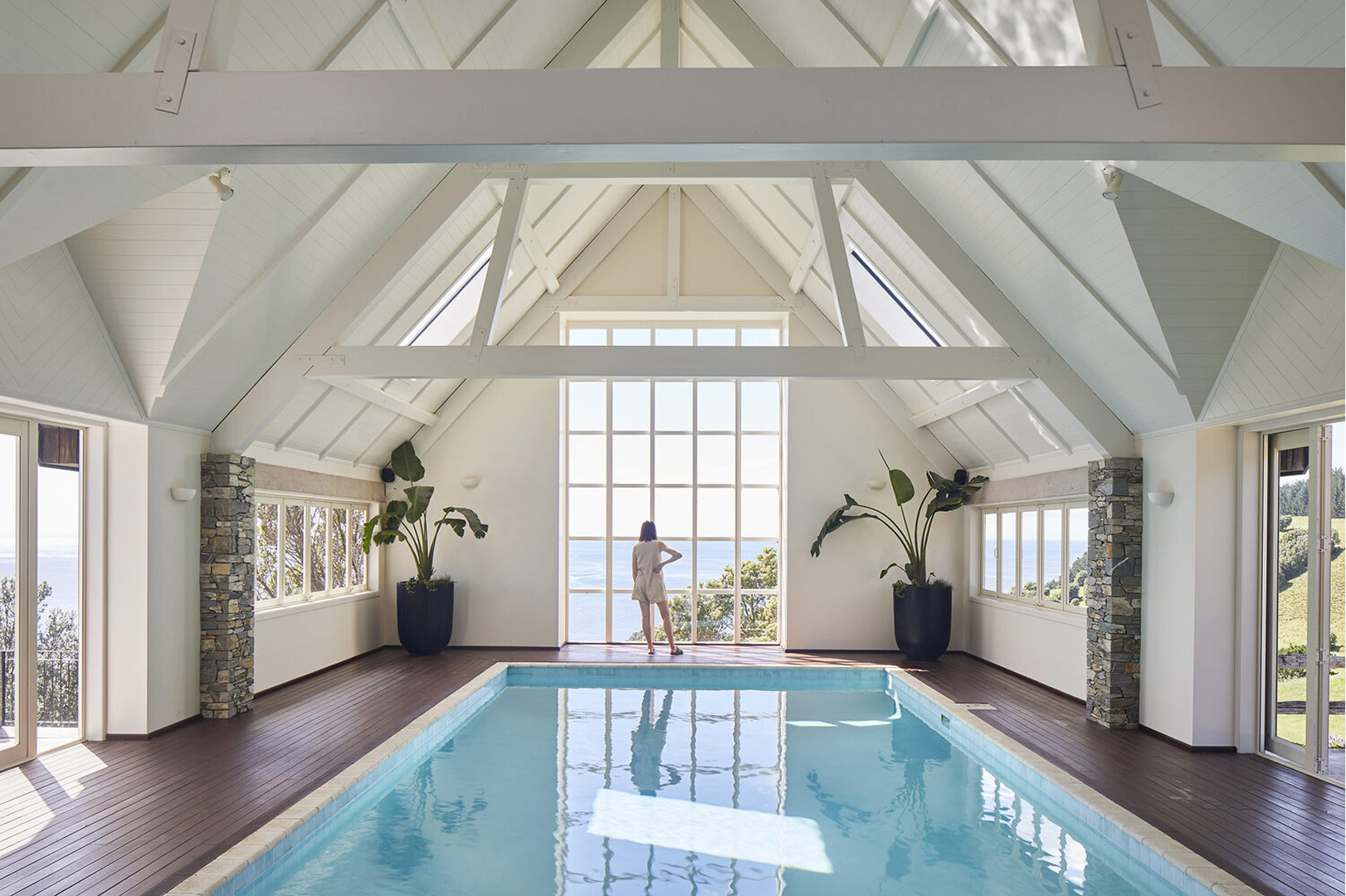 Indoor pool room with high white ceiling, large window, two plants, and a person looking outside.