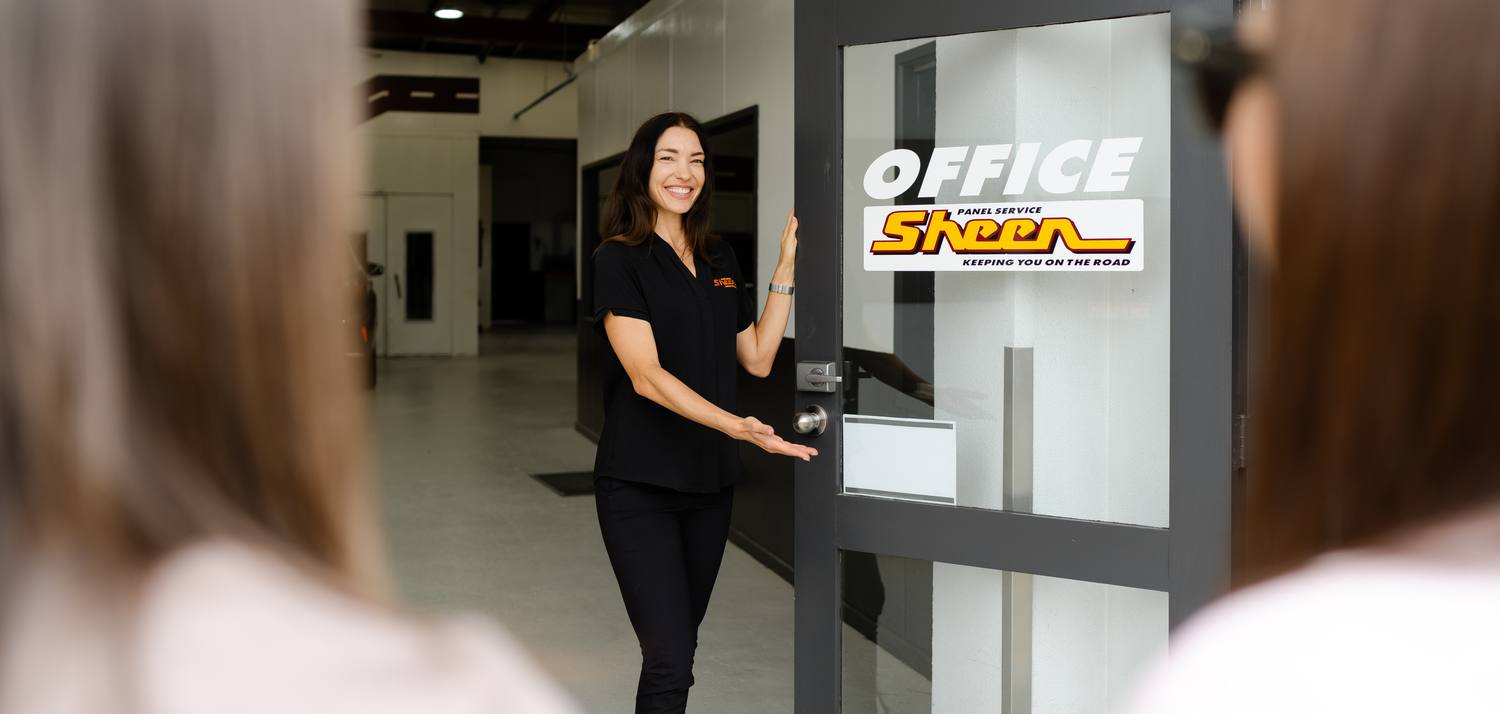 Smiling woman in black uniform holds open a glass door with Sheen Panel Service office signage.