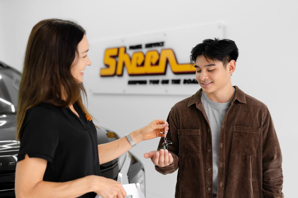 Woman handing car key to smiling man in front of vehicle and Sheen Motors sign.