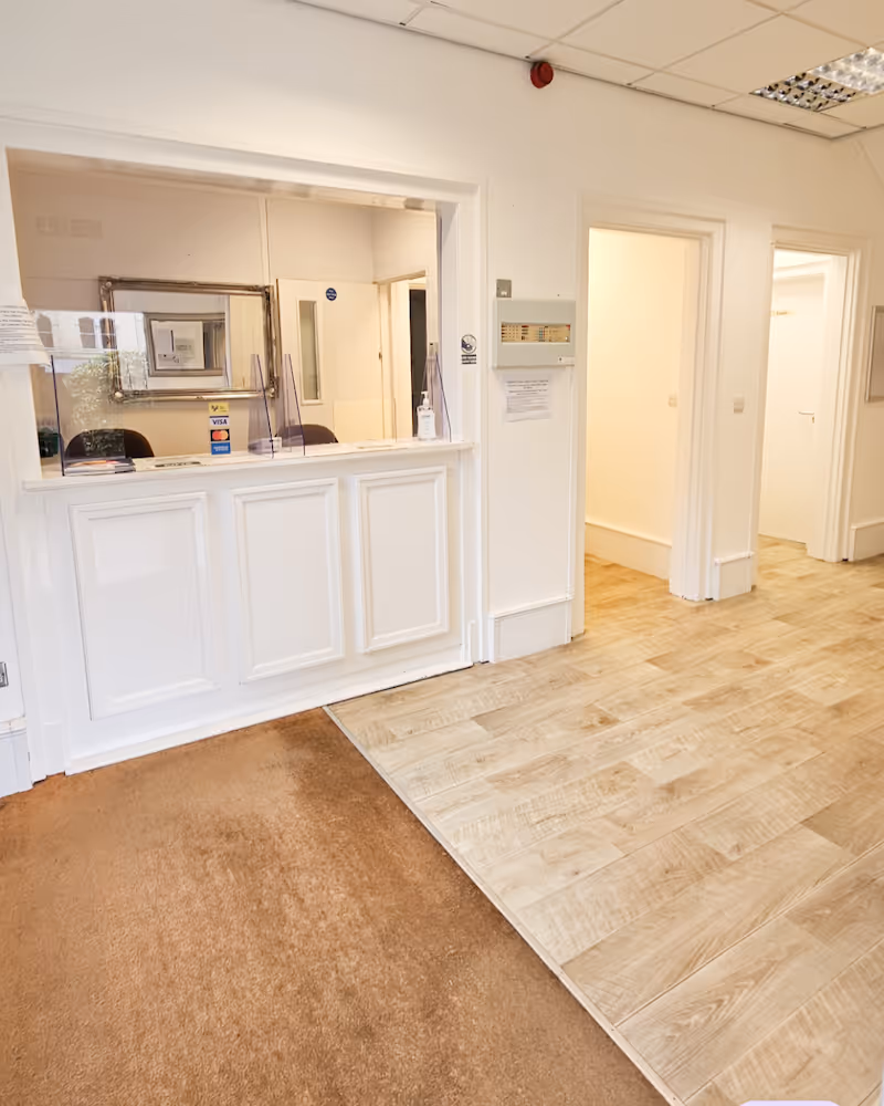 Bright office reception area with a white counter and glass window, two doorways leading to other rooms, and wood-patterned floor alongside brown carpet.