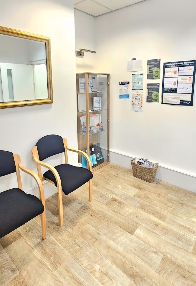 Waiting area with two black cushioned wooden chairs, a glass display cabinet with informational brochures, a large wall mirror, and a basket of magazines on the wooden floor.