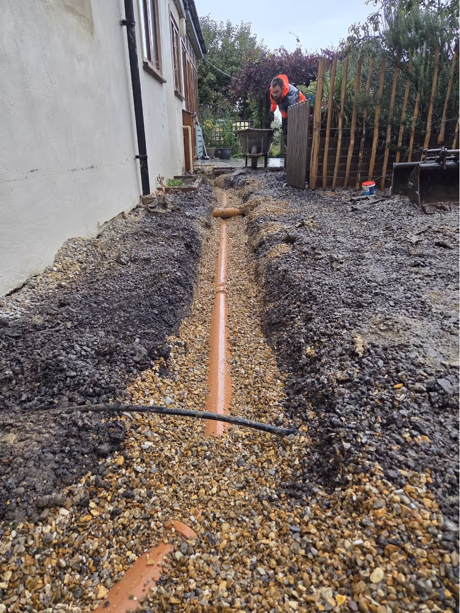 Person working on a trench with an exposed pipe running alongside a house.