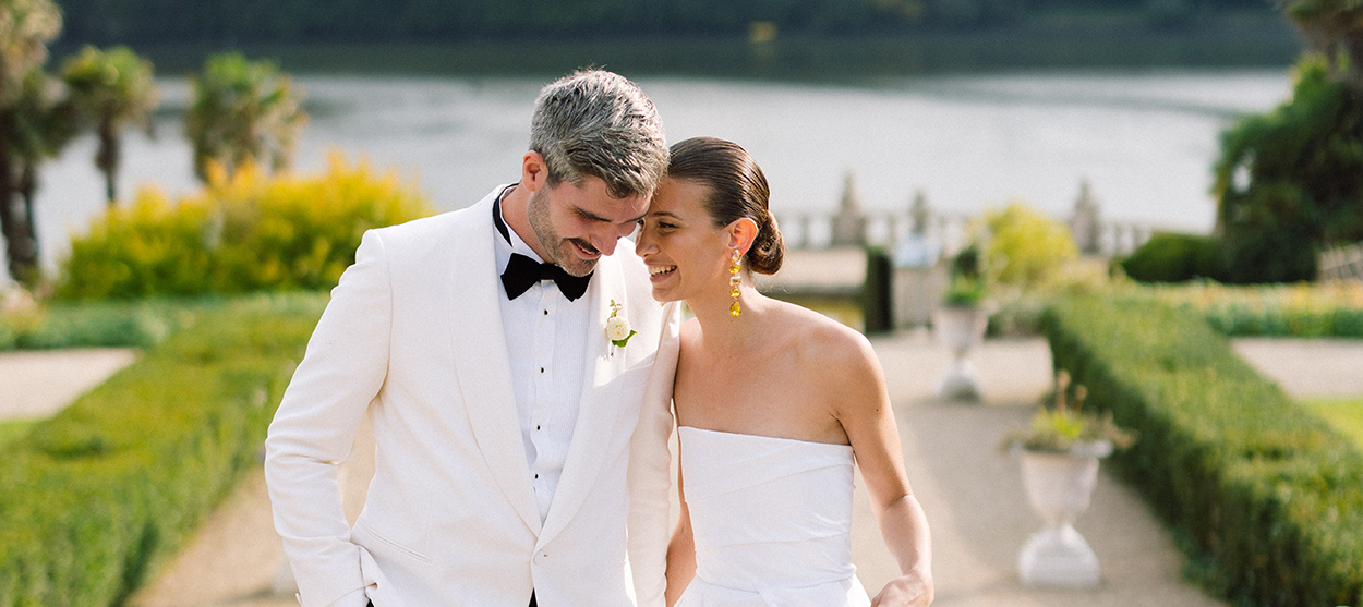 Smiling couple in white attire walking closely together outdoors near a lake with greenery and decorative urns.