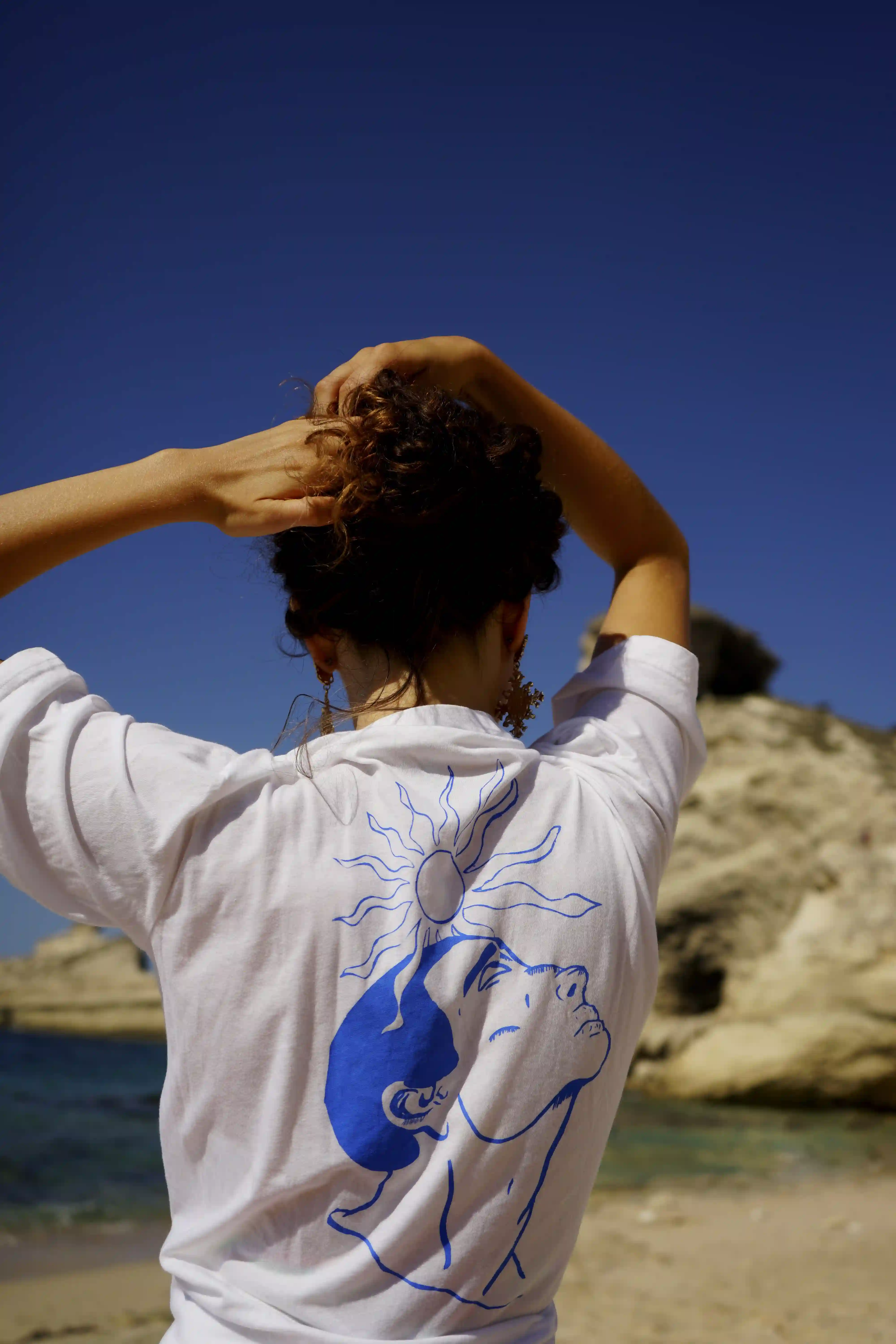 Photographie de mode montrant une femme de dos soulevant ses cheveux pour dévoiler une sérigraphie bleue sur un t-shirt blanc, face à la baie de la Pointe Saint-Antoine à Bonifacio, Corse.