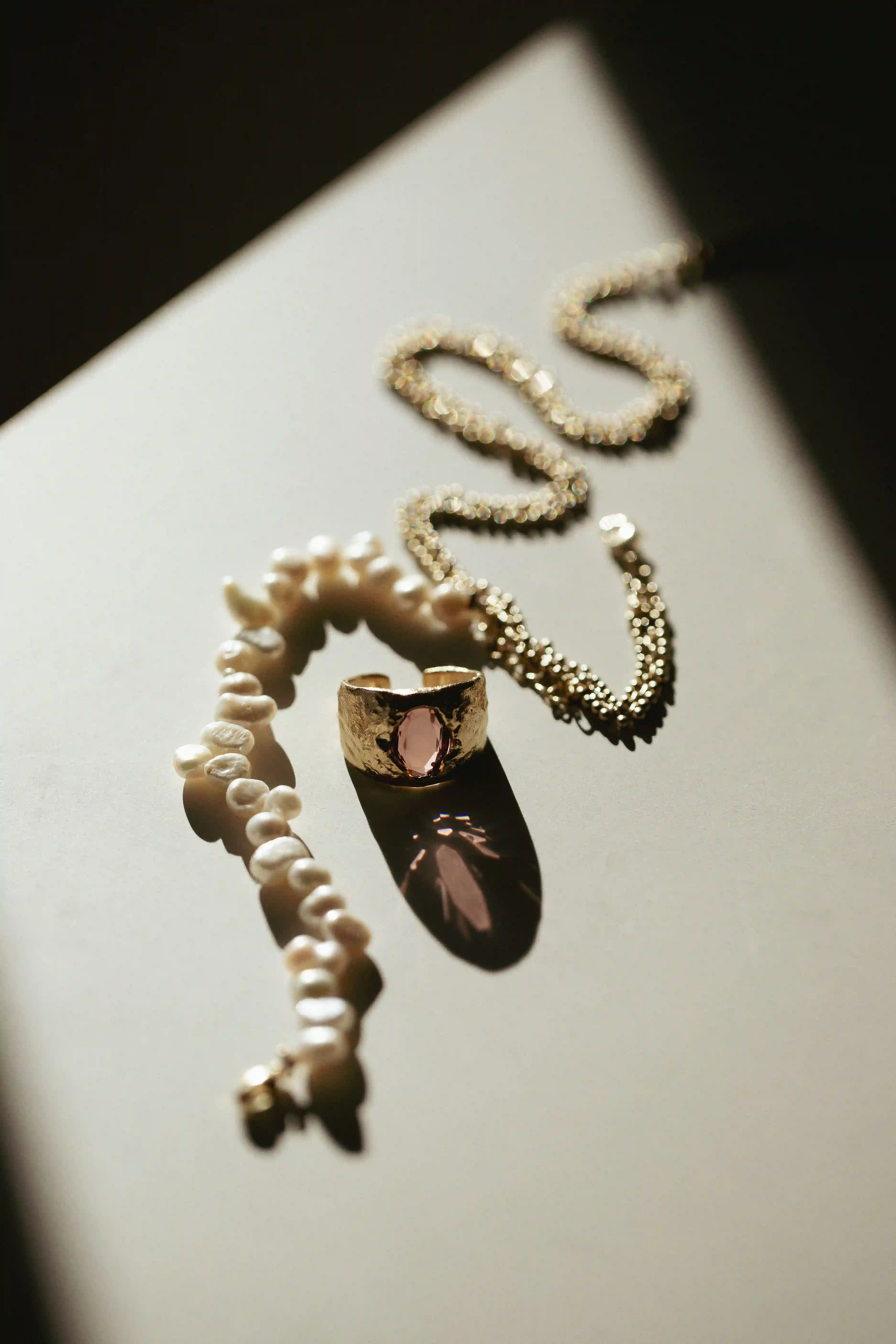Photographie de bijoux : bague en or avec pierre rose, collier et bracelet en or posés sur fond blanc, éclairés par un contre-jour solaire.