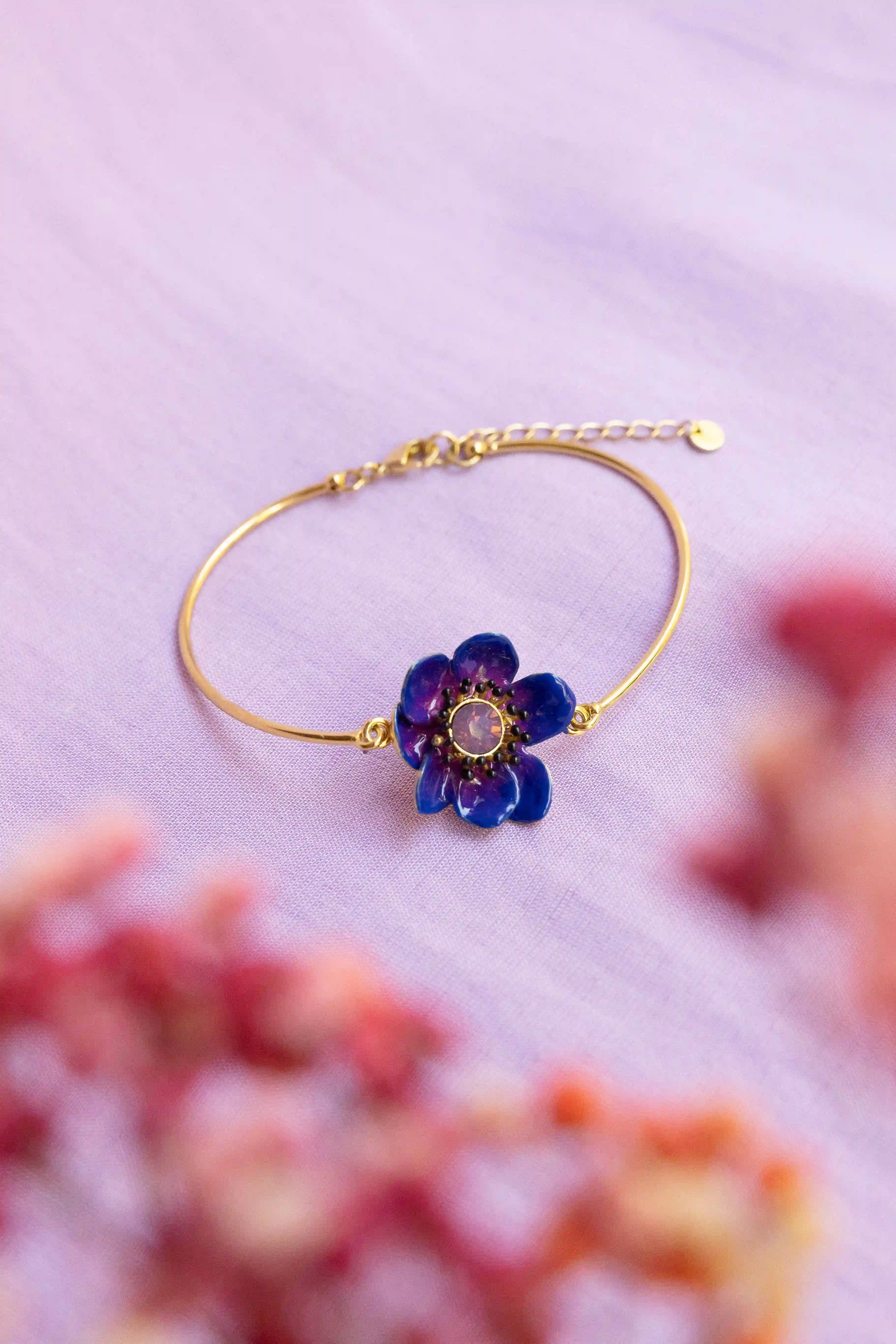 Jonc en or orné d’une fleur violette, photographié sur un fond lilas avec des fleurs roses au premier plan.