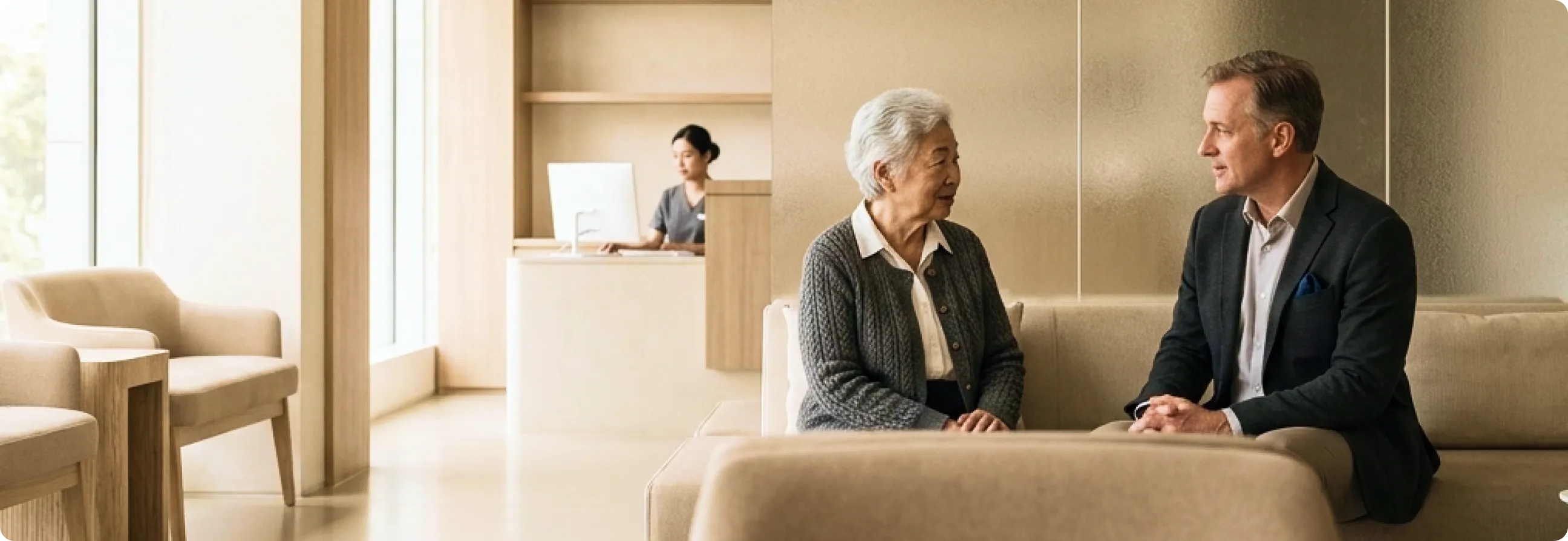 An elderly woman and a middle-aged man sitting and talking on a beige sofa in a modern, warmly lit waiting area with a receptionist working at a desk in the background.