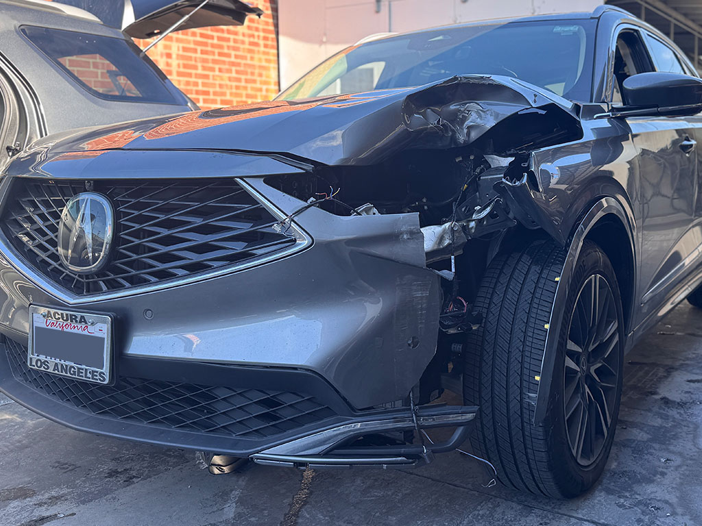 Front-end collision damage on a grey Acura MDX featuring crushed hood, broken headlight, and exposed structural components