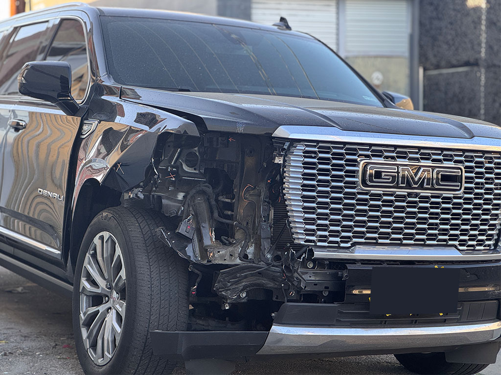 Black GMC Yukon Denali with severe passenger-side front-end collision damage, including missing headlight assembly, crushed fender, and exposed internal cooling components.
