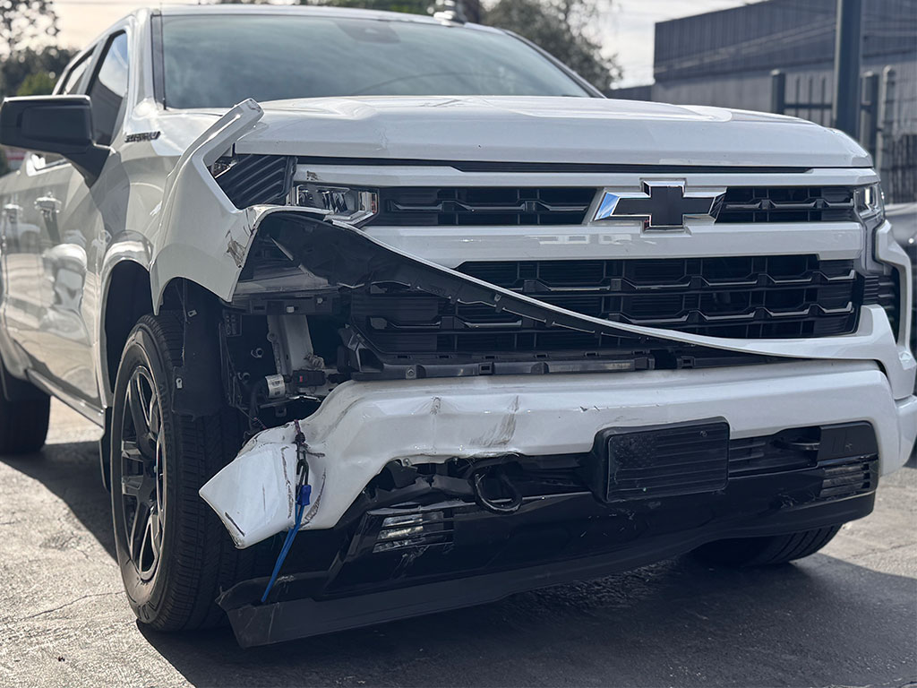 Low-angle shot of a damaged white Chevrolet Silverado’s front passenger-side bumper area with severe crush damage, torn fascia, and crumpled structural metal.