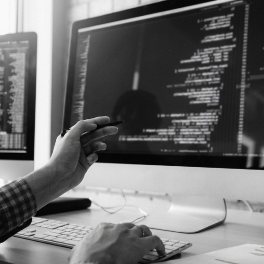 Person pointing with pen at code displayed on a computer monitor while typing on keyboard.
