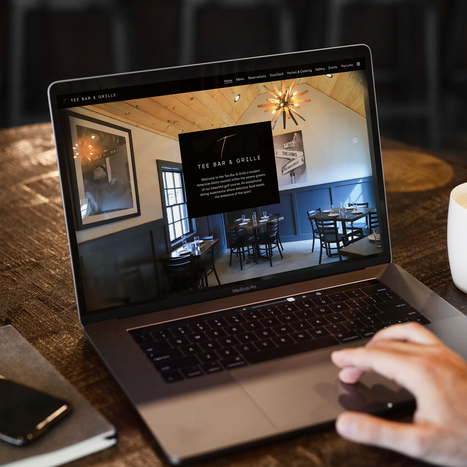 Person using a MacBook Pro displaying the Tee Bar & Grille restaurant website with an interior photo of the dining area.