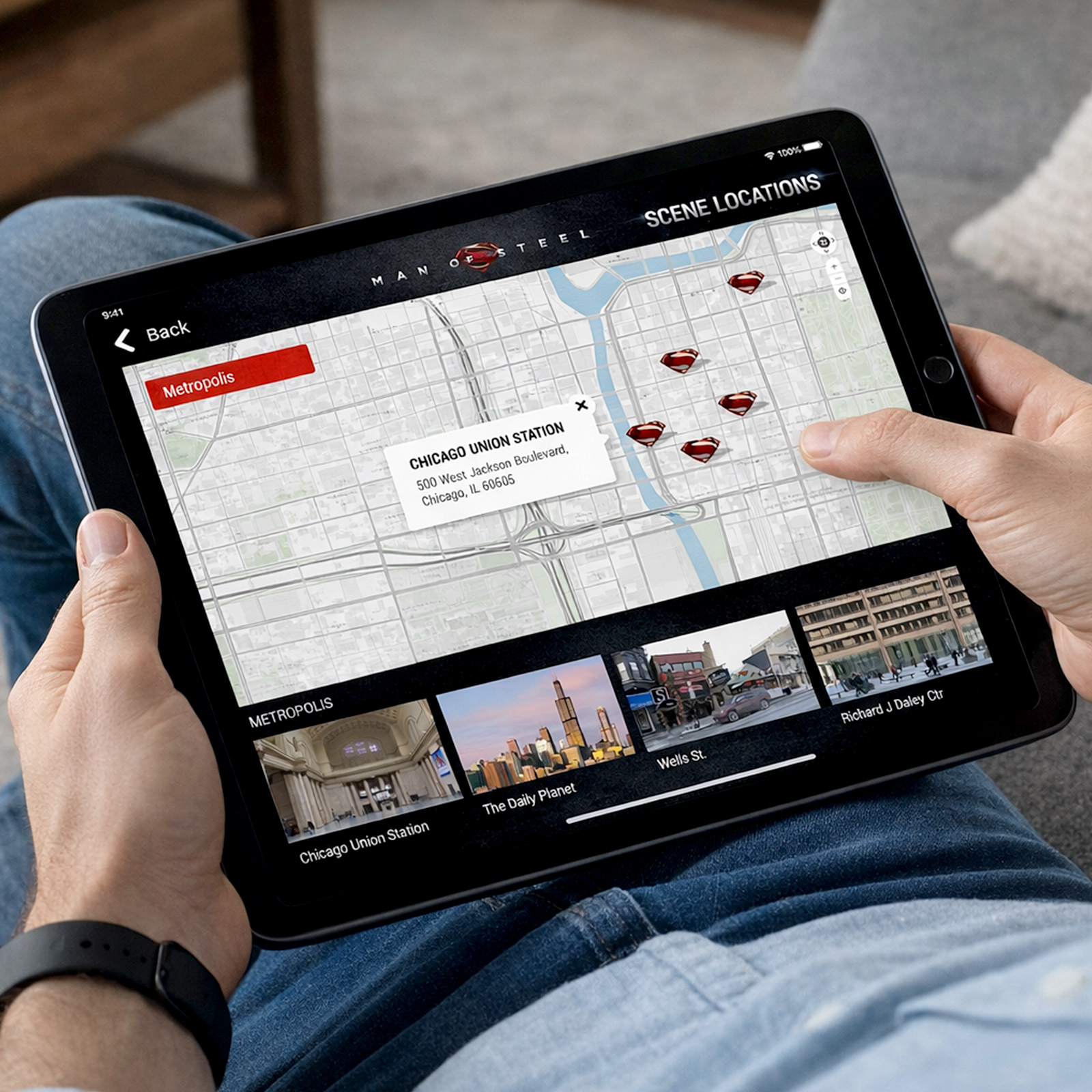 Person holding a tablet showing a Man of Steel scene locations map of Metropolis with marked sites and thumbnails including Chicago Union Station and The Daily Planet.