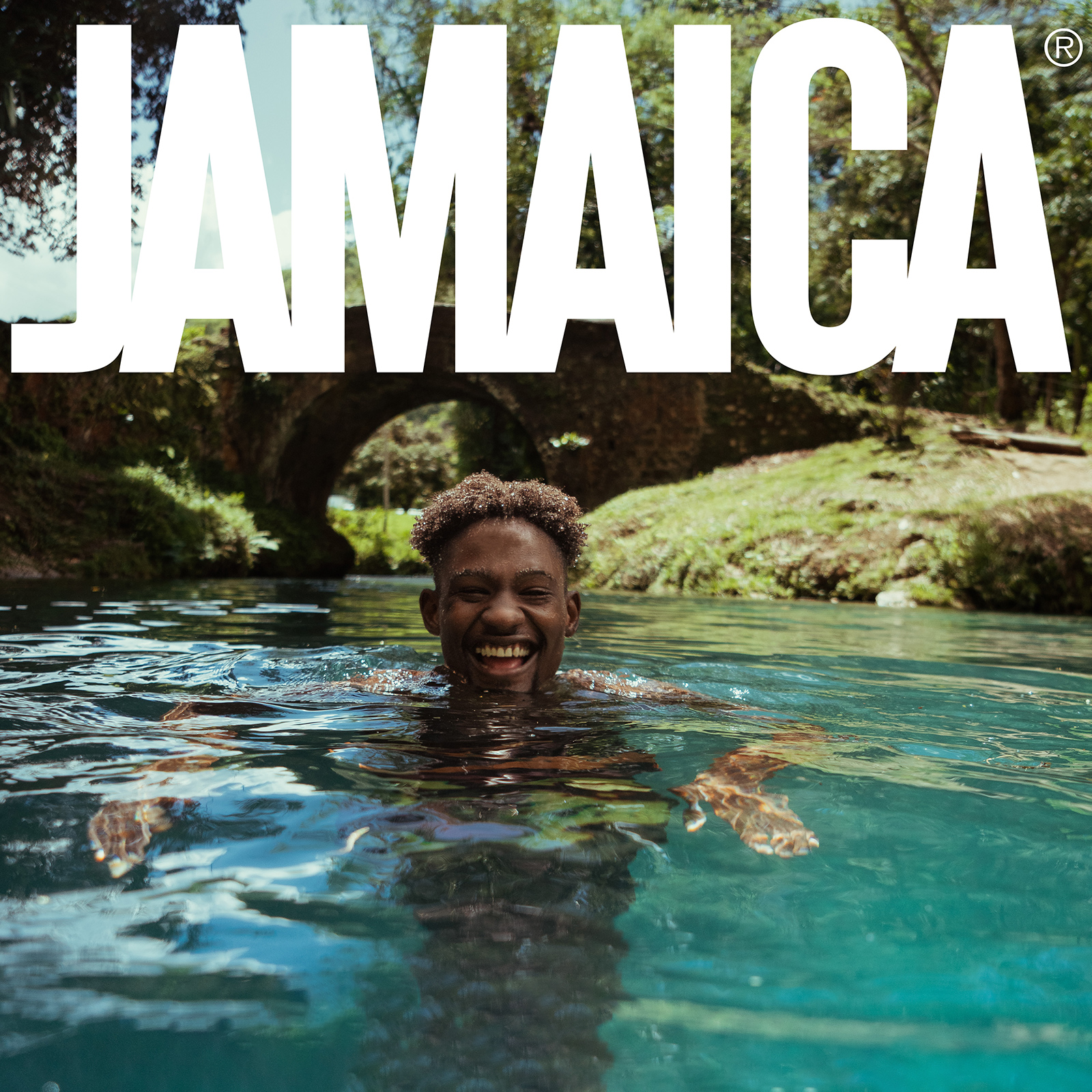 Smiling man swimming in clear turquoise water with a stone arch bridge and lush greenery in the background, overlaid with large text 'JAMAICA'.