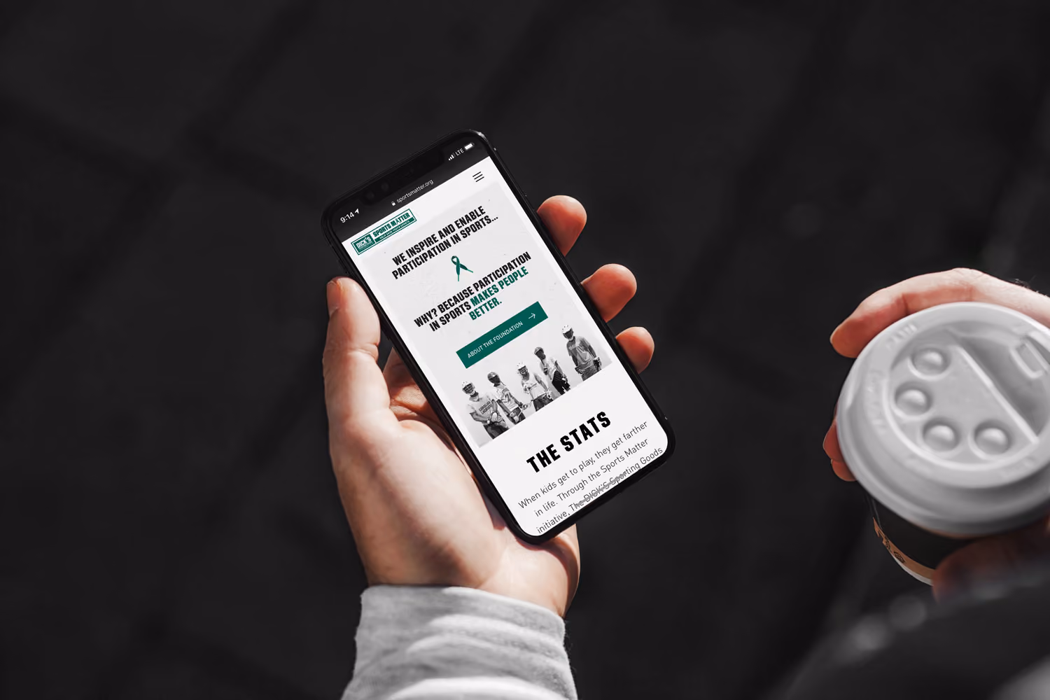 Person holding smartphone displaying sports participation website and coffee cup with lid.