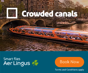 Tour boat with passengers passes under a stone bridge on a crowded canal in an urban setting.
