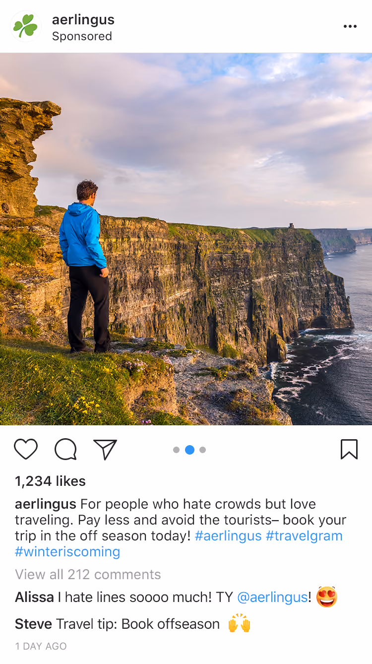 Man in blue jacket stands on green cliff edge overlooking tall cliffs and ocean under cloudy sky.