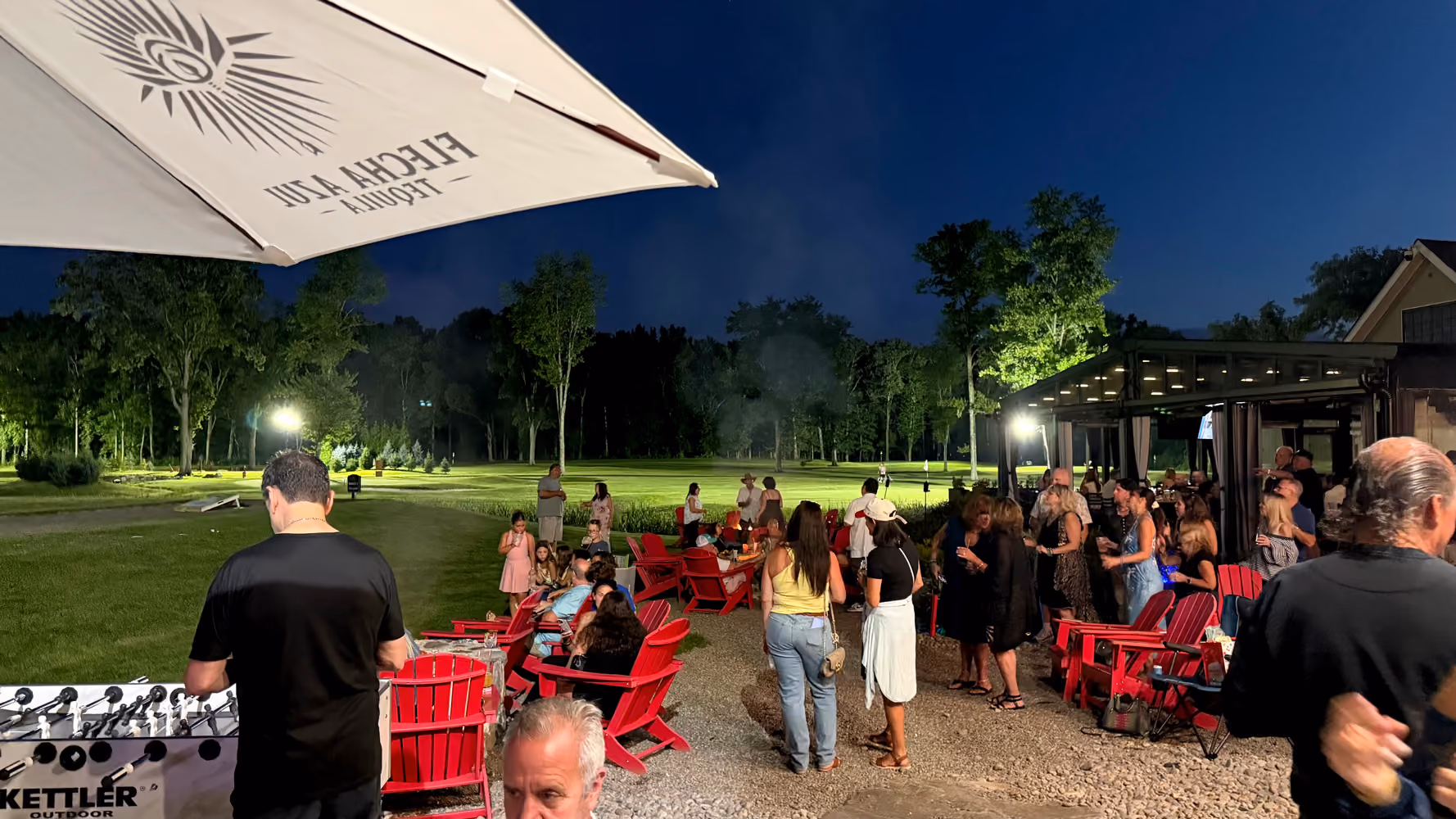 People socializing outdoors at night with red chairs, a foosball table, and umbrella on a lawn.
