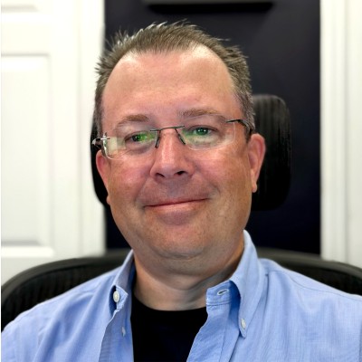 Middle-aged man with glasses smiling, wearing a blue button-up shirt over a black top, seated indoors.