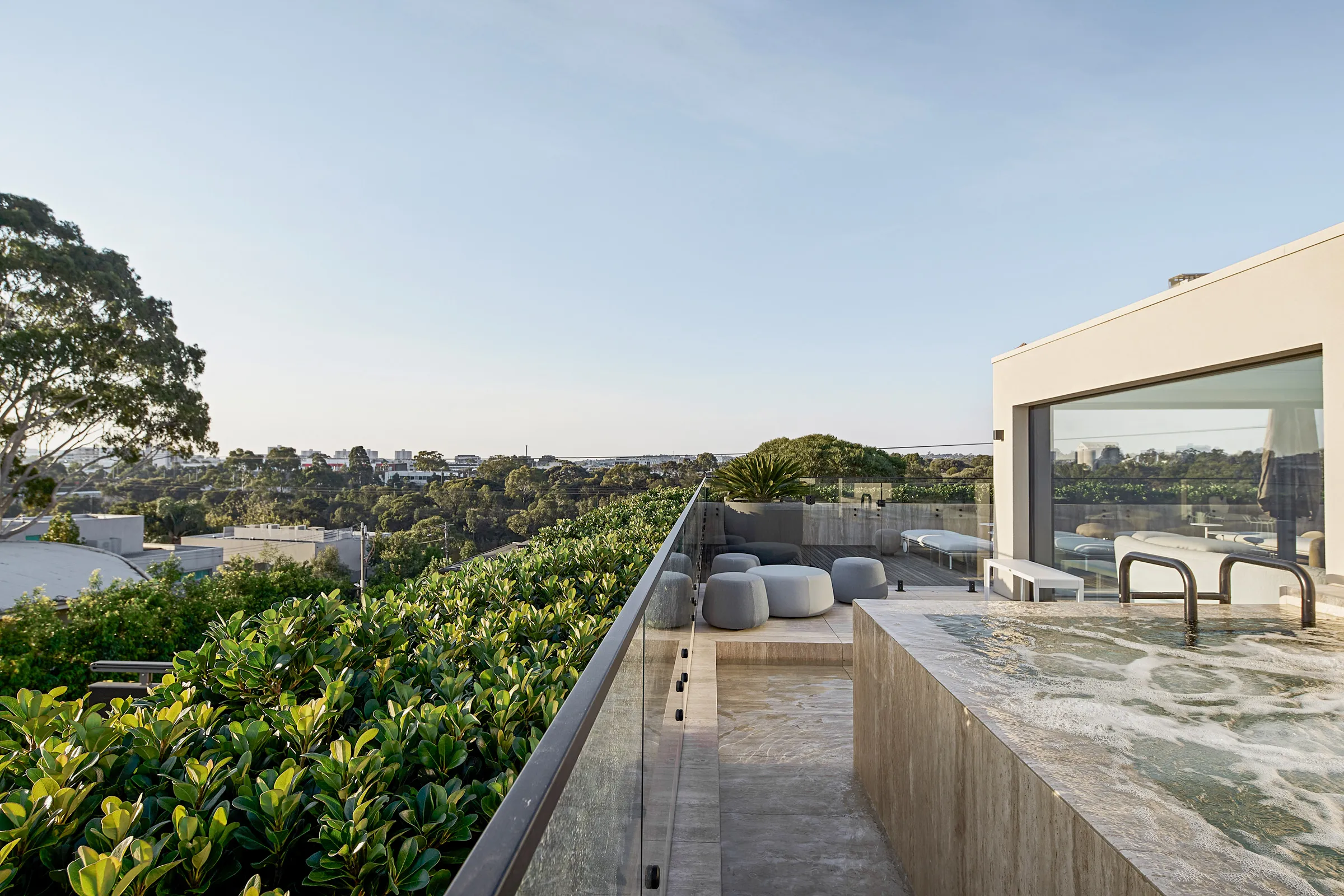Modern rooftop terrace with bubbling hot tub, gray cushioned seating, glass railing, and greenery overlooking a tree-lined cityscape.