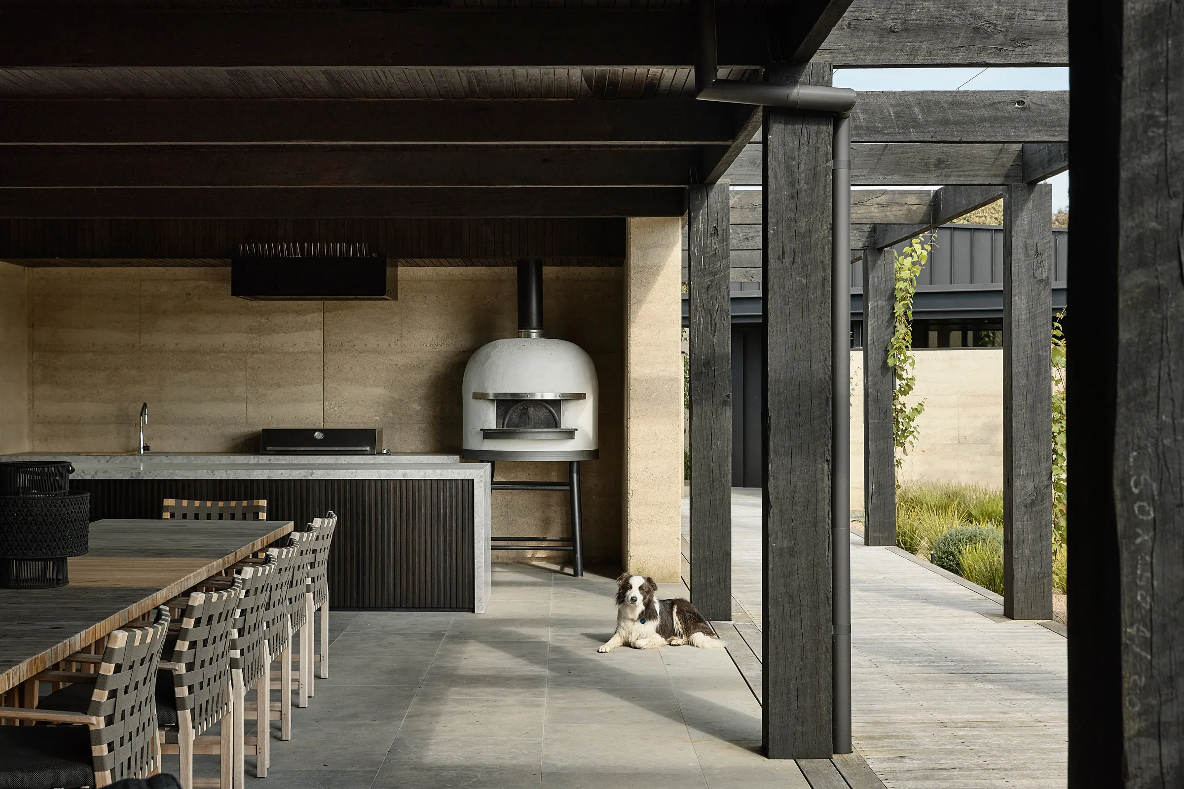 Modern outdoor kitchen with wooden dining table and chairs, white pizza oven, and a dog lying on the tiled floor.