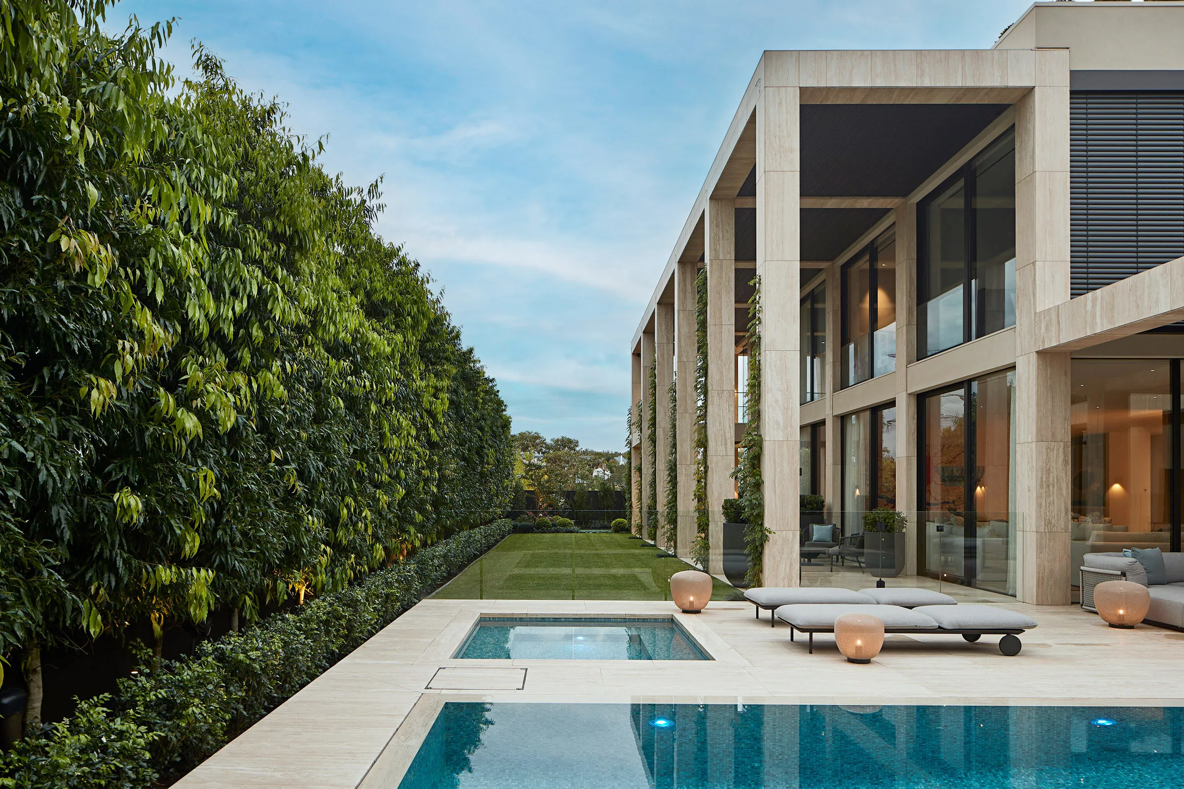 Modern house with tall beige columns, glass walls, lounge chairs, pool, and a tall green hedge along the side garden.