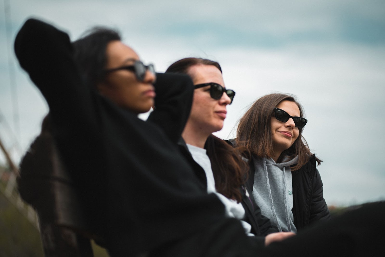 Three researchers wearing sunglasses. The first is leaning back, with her arms crossed behind her back, looking thoughtful but neutral. Th second one has a faint smile, but her position is less relaxed. The third one has a wide smile, slouching comfortably.