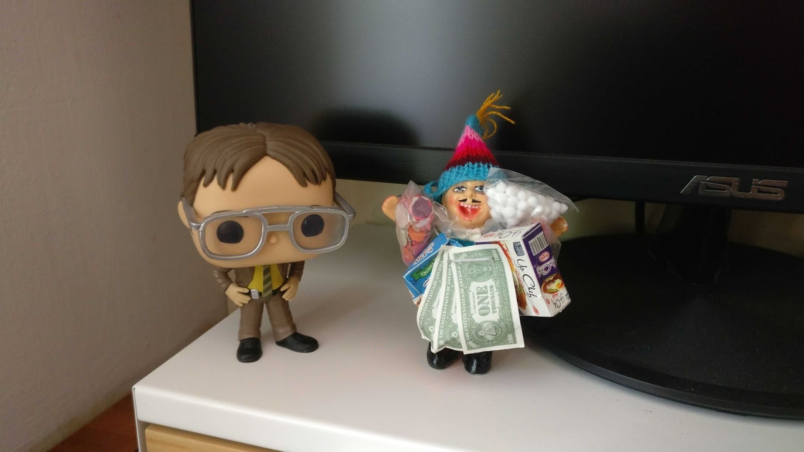 A picture of two small dolls standing in front of a screen. A Dwight Schrute's Funko and a Bolivian Ekeko.