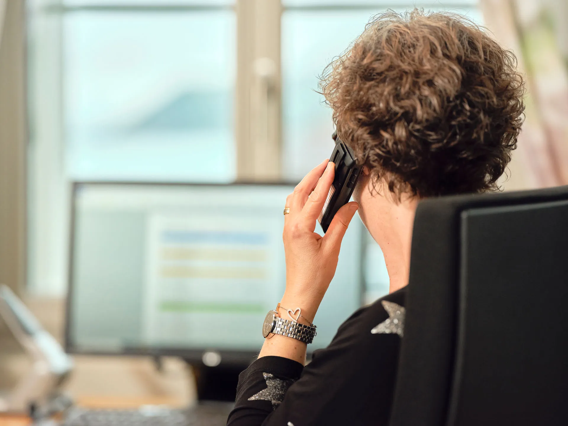 Frau mit lockigem Haar telefoniert am Arbeitsplatz vor einem verschwommenen Computermonitor.