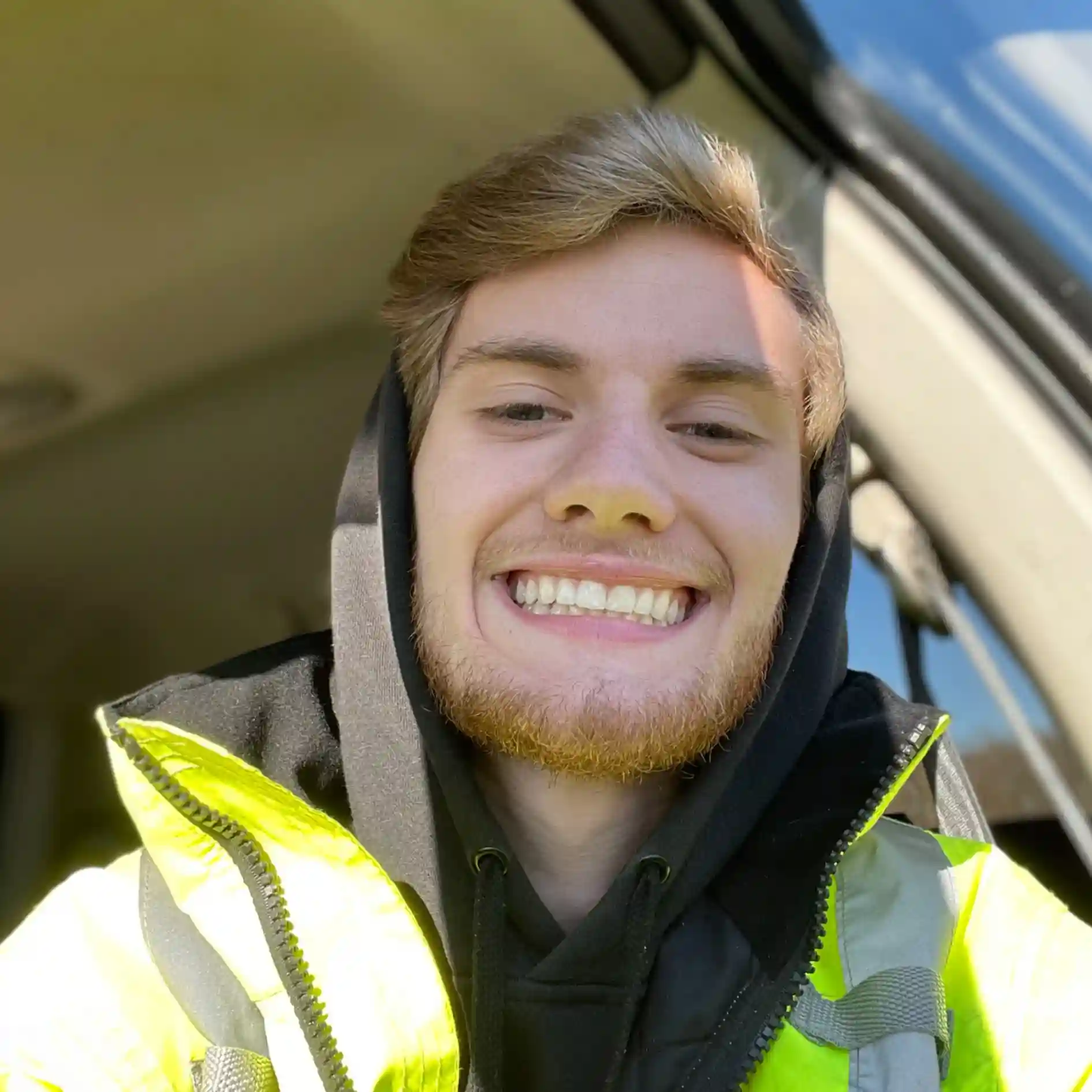 Smiling young man with light brown hair and beard wearing a black hoodie and a yellow high-visibility jacket inside a vehicle. Selfie