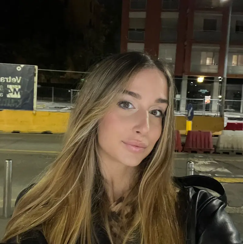 Young woman with long blonde hair taking a selfie outdoors at night near a street with barriers and a building in the background.