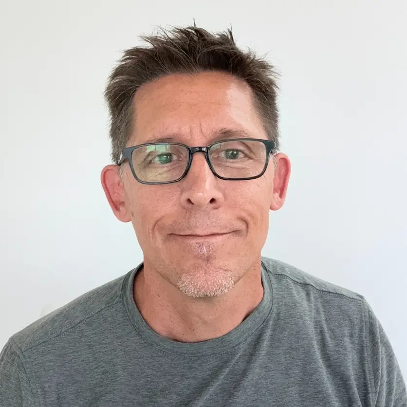 Man with short brown hair, glasses, and gray t-shirt smiling gently against a plain white background. Made with phone 