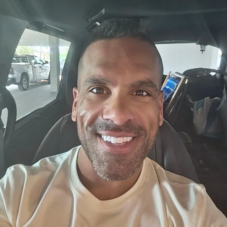 Smiling man with short hair and beard sitting inside a car wearing a light-colored shirt. Selfie taken before using AI SuitUp