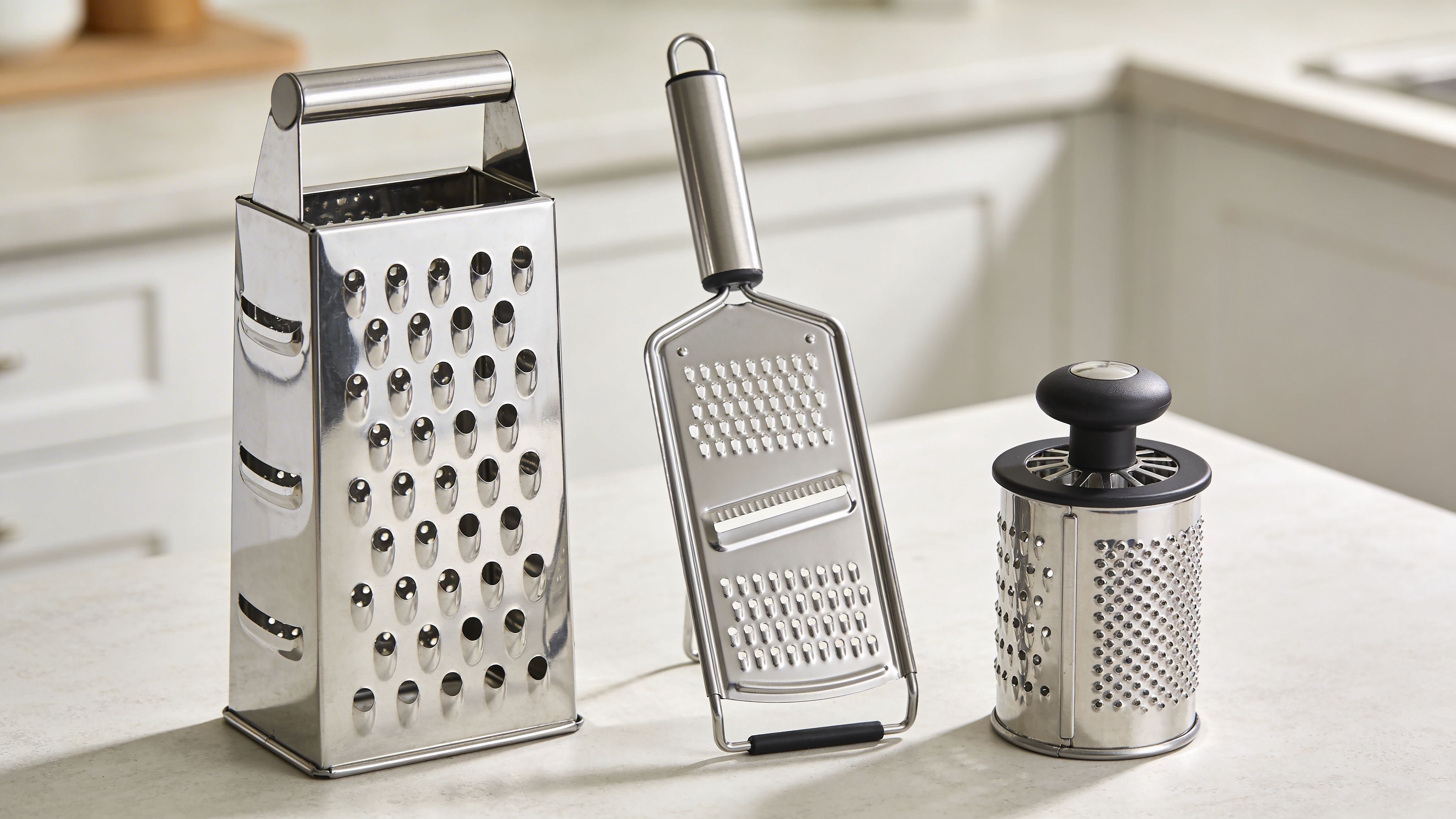 Three different styles of stainless steel kitchen graters sitting on a white marble countertop.