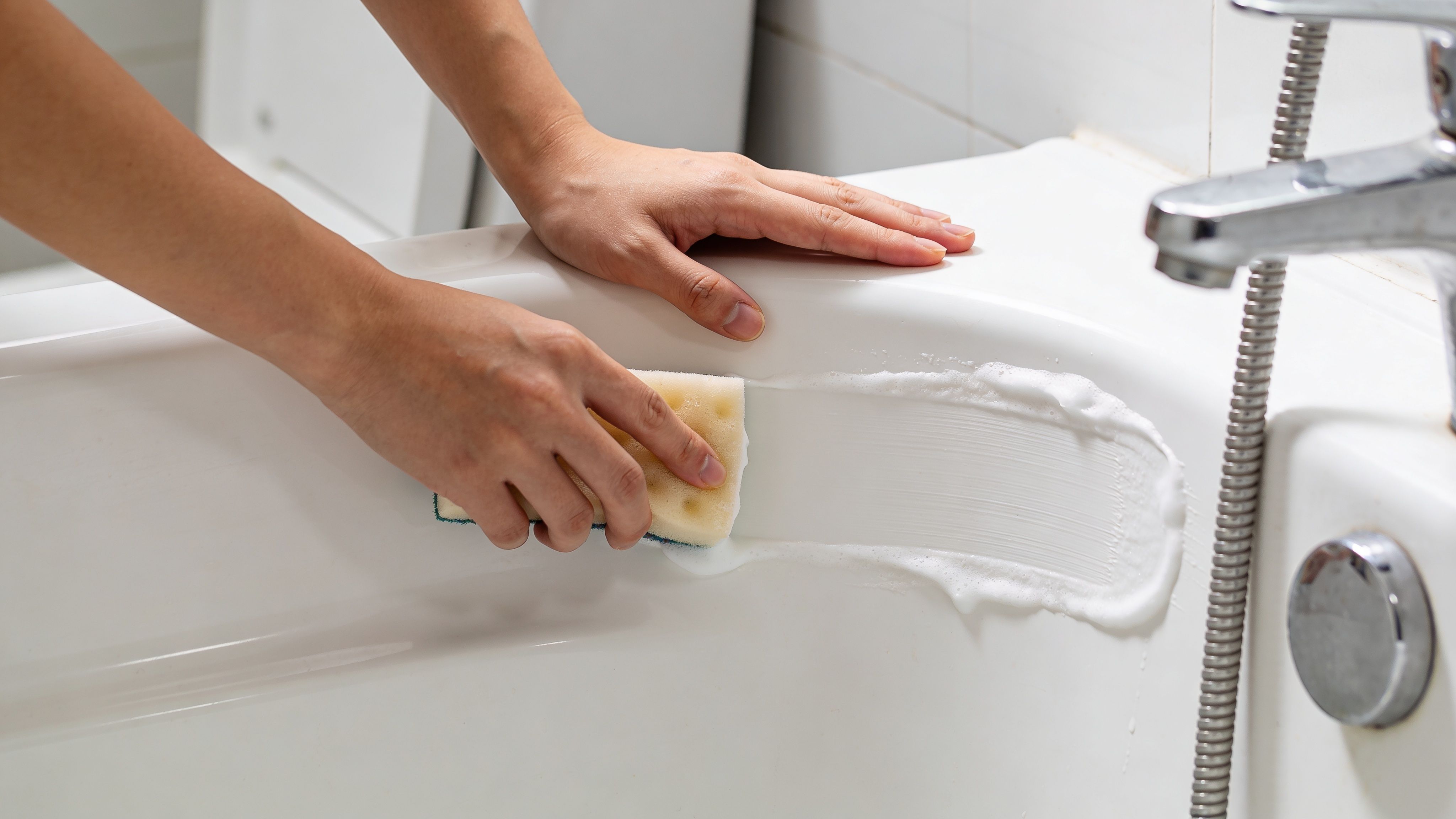 A person using a yellow sponge to scrub a white plastic bathtub clean with soap suds.