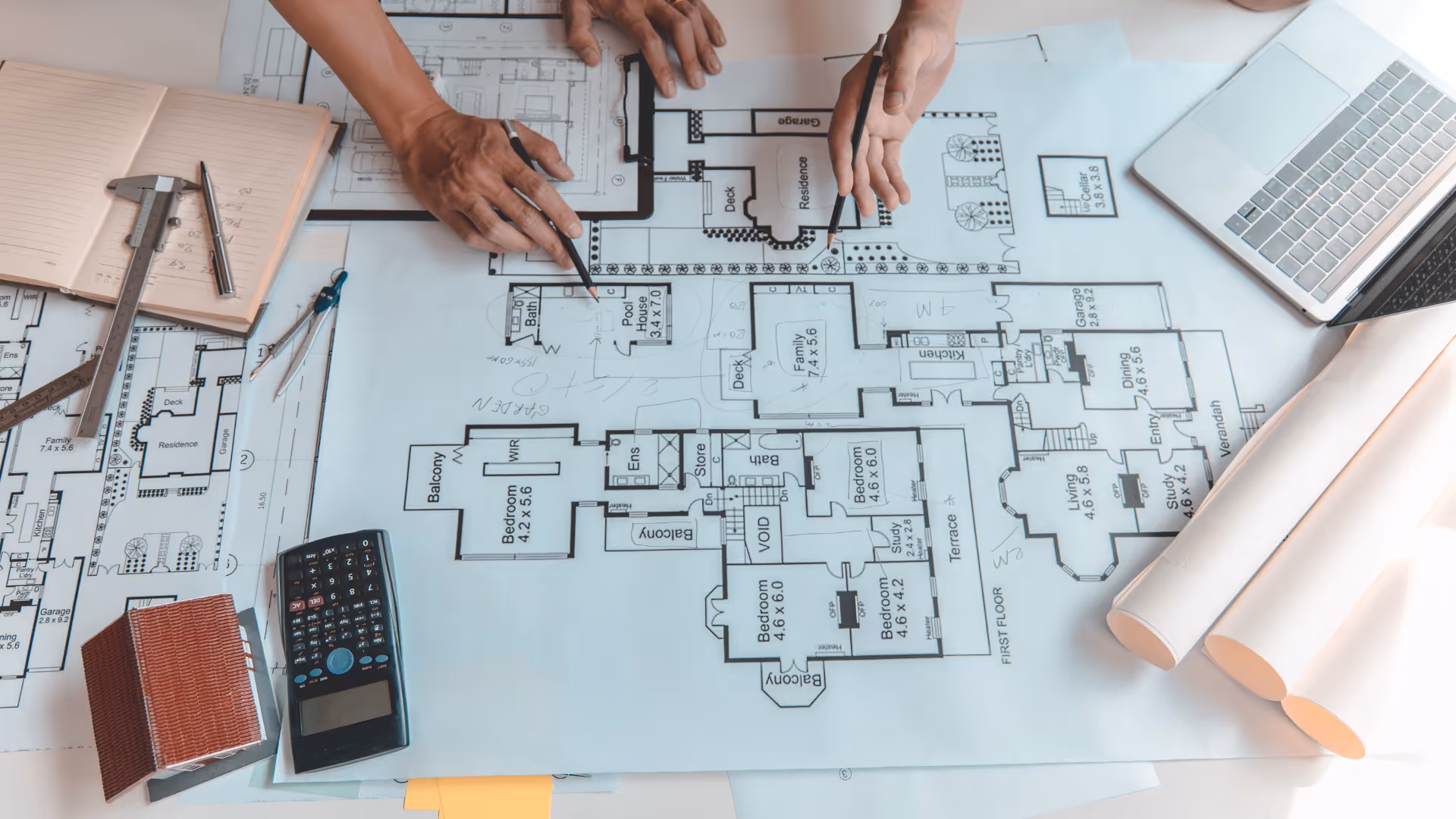 Architekten besprechen Baupläne an einem Tisch mit technischen Zeichnungen, Lineal, Taschenrechner, Notizbuch, Laptop und Modellhaus – symbolisiert Planungsphase eines Bauprojekts.