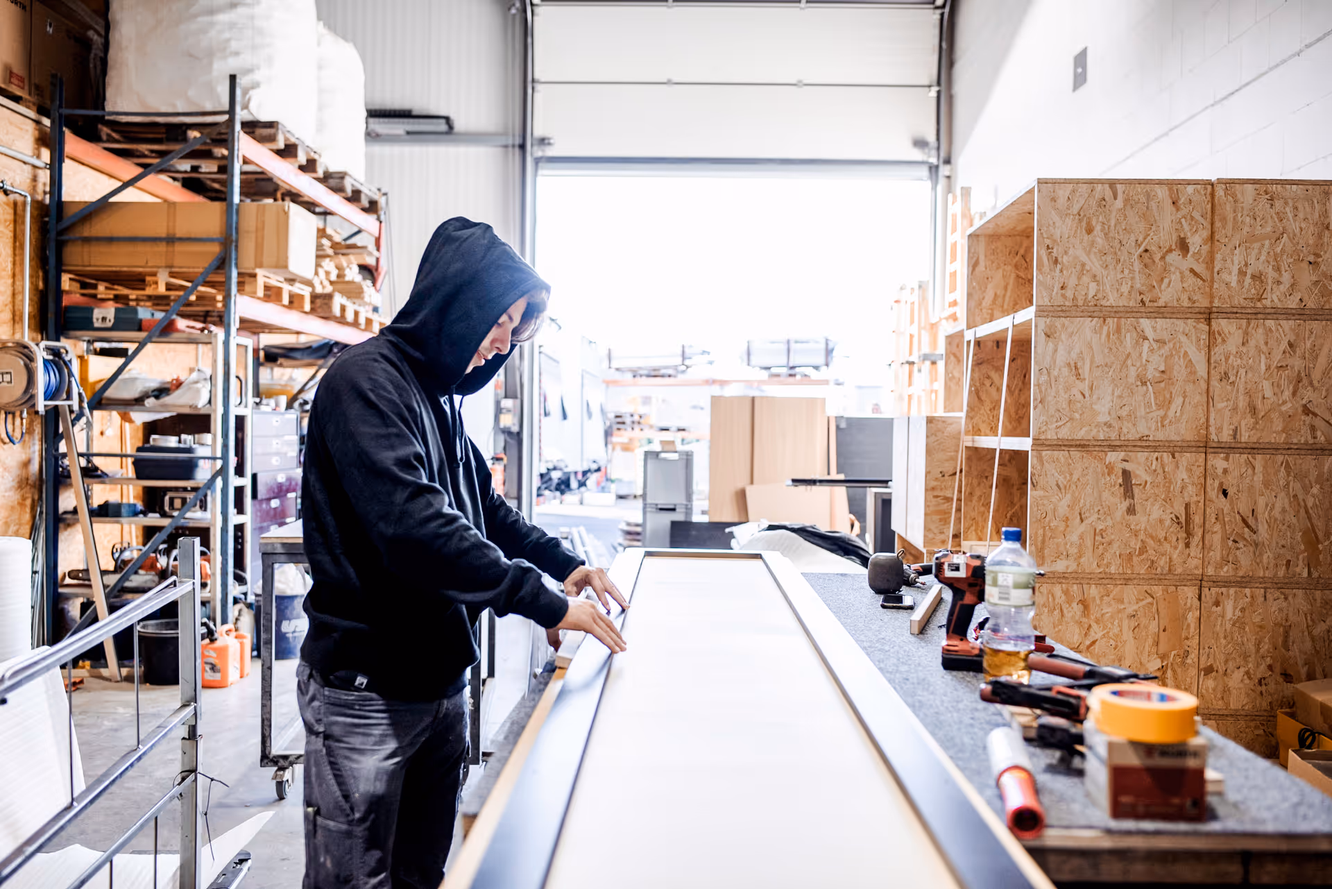 Tischler bei der Arbeit in einer Werkstatt: Eine Person in Arbeitskleidung und Kapuzenpullover richtet ein Werkstück aus, umgeben von Werkzeug, Holzplatten und Regalen – typische Szene aus dem Produktionsalltag in einem Handwerksbetrieb.