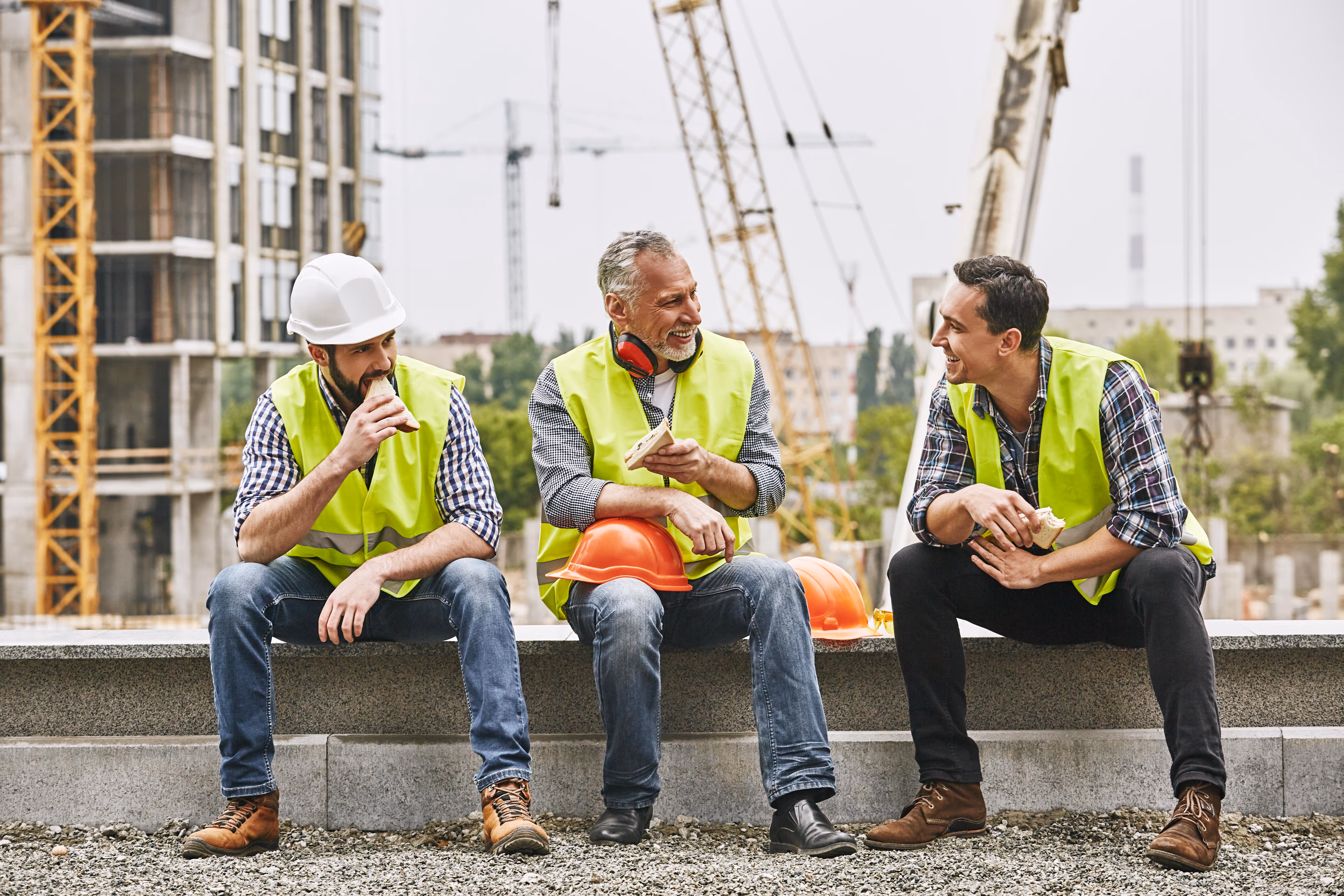 Drei Bauarbeiter in Warnwesten machen auf einer Baustelle Pause. Sie sitzen lachend nebeneinander auf einer Betonkante, essen belegte Brote und unterhalten sich. Im Hintergrund sind Kräne, ein Rohbau und weitere Bauarbeiten zu sehen.