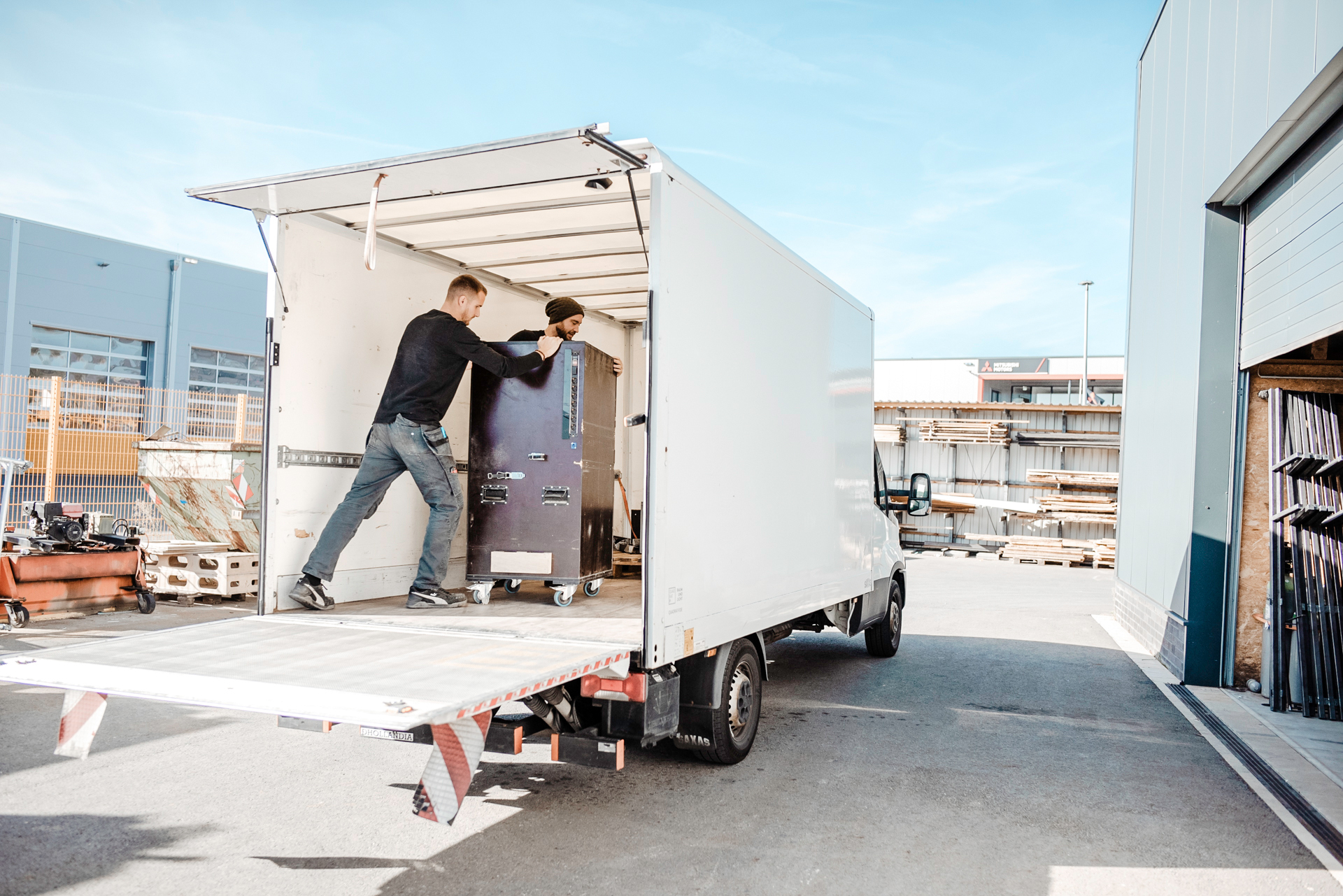 Zwei Männer laden einen großen Metallschrank mit Rollen auf einen Transporter im Industriehof